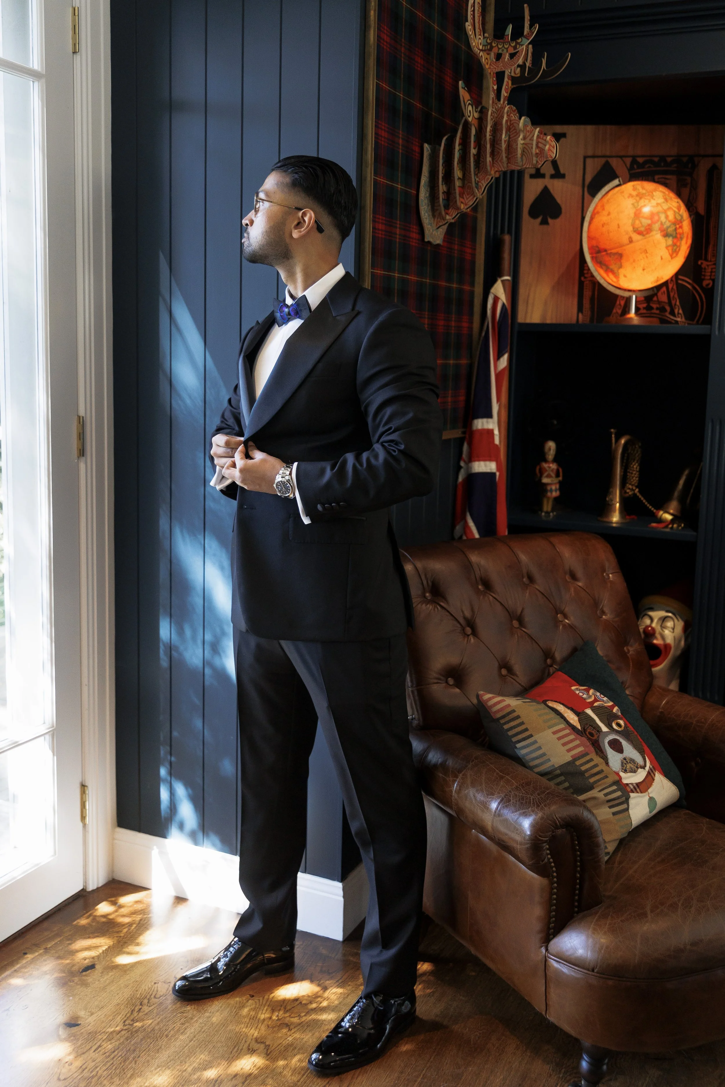 A man in a black tuxedo with a blue bow tie adjusting his jacket indoors, near a window with sunlight, in a room decorated with Christmas and British-themed items, including a Union Jack flag, a stag head, and various decorative objects.