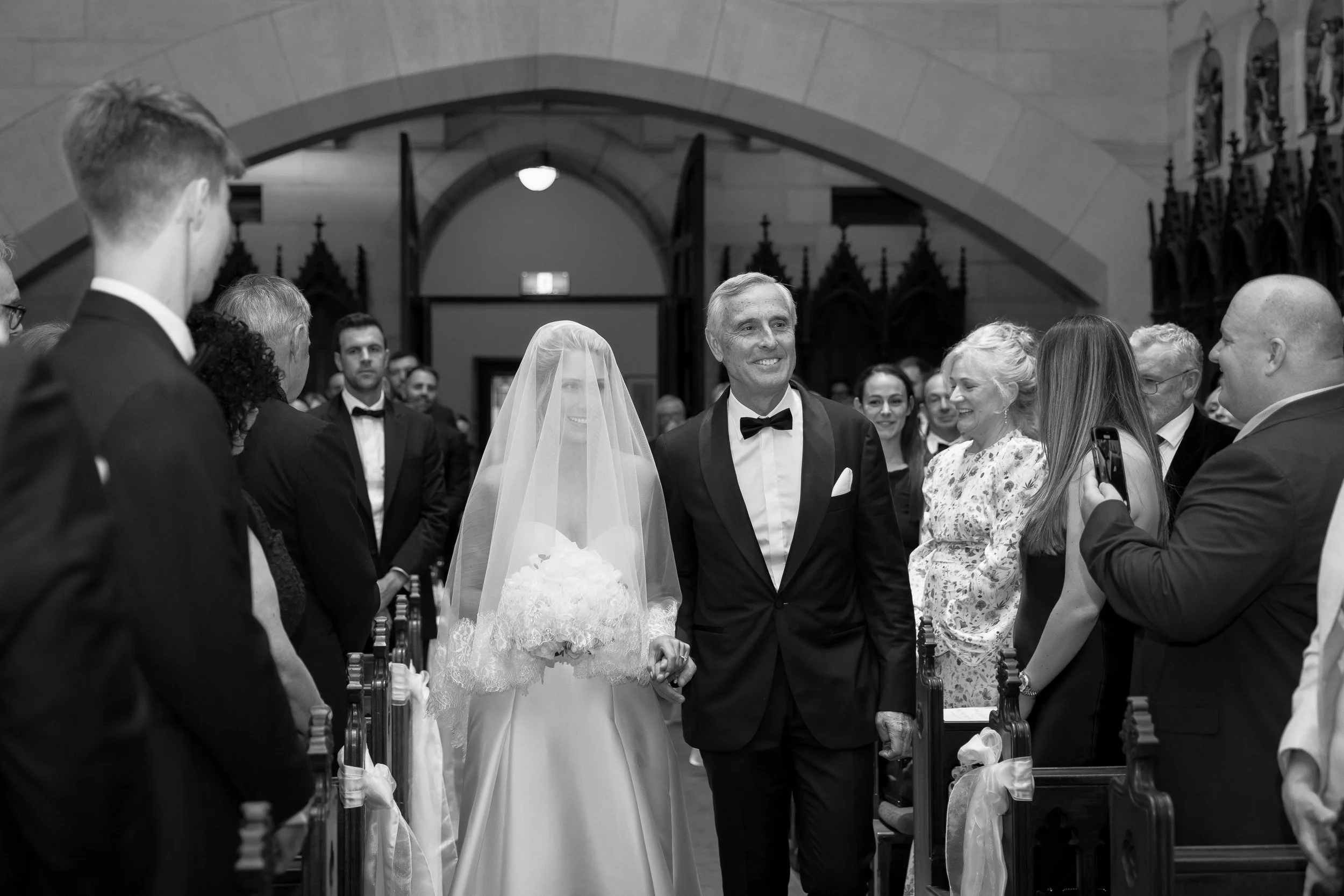 A bride and her father walking down the aisle during a wedding ceremony inside a church, surrounded by guests dressed in formal attire.