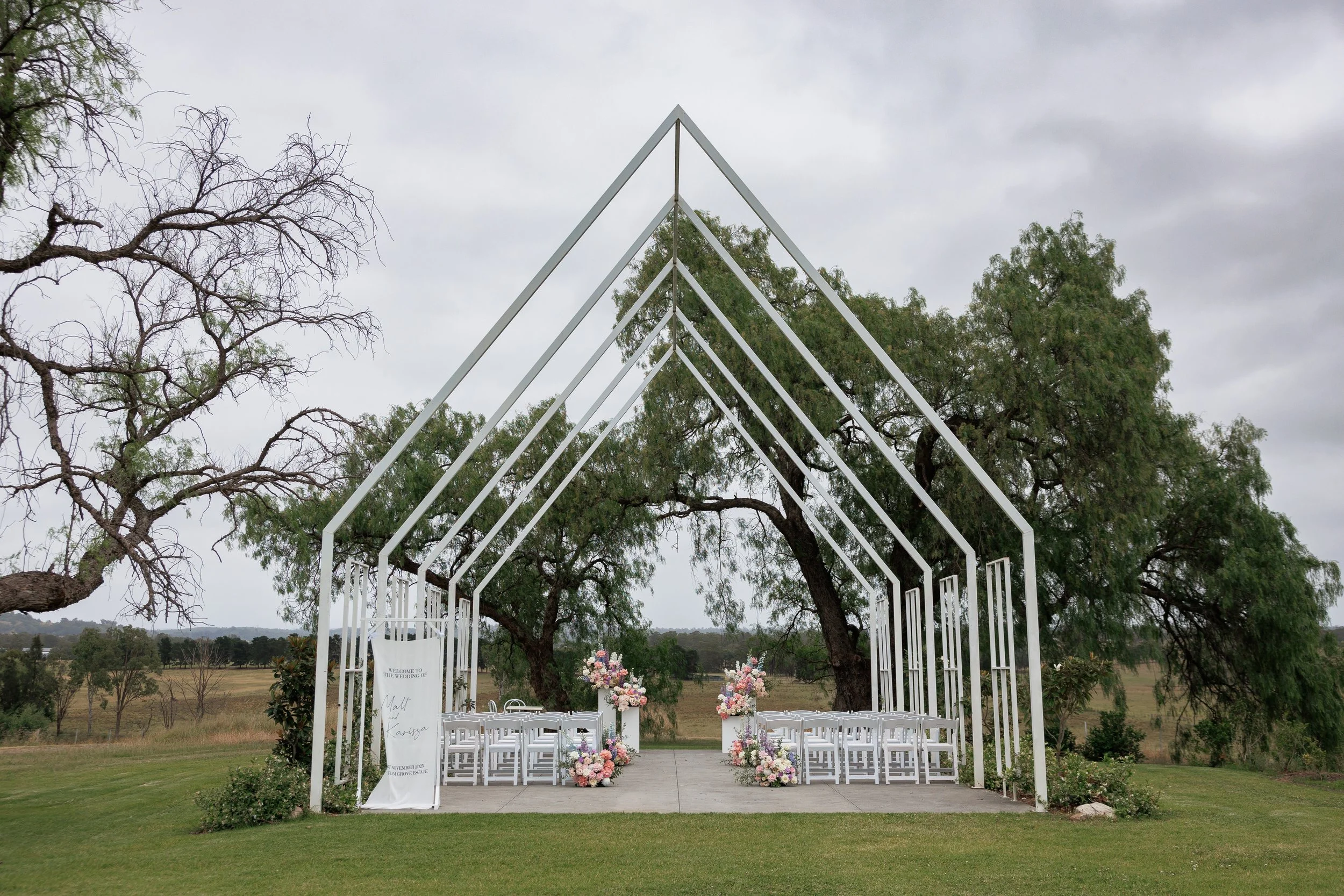 Outdoor wedding altar with white metal arch structure, decorated with floral arrangements, surrounded by green trees, on a grassy lawn under a cloudy sky.