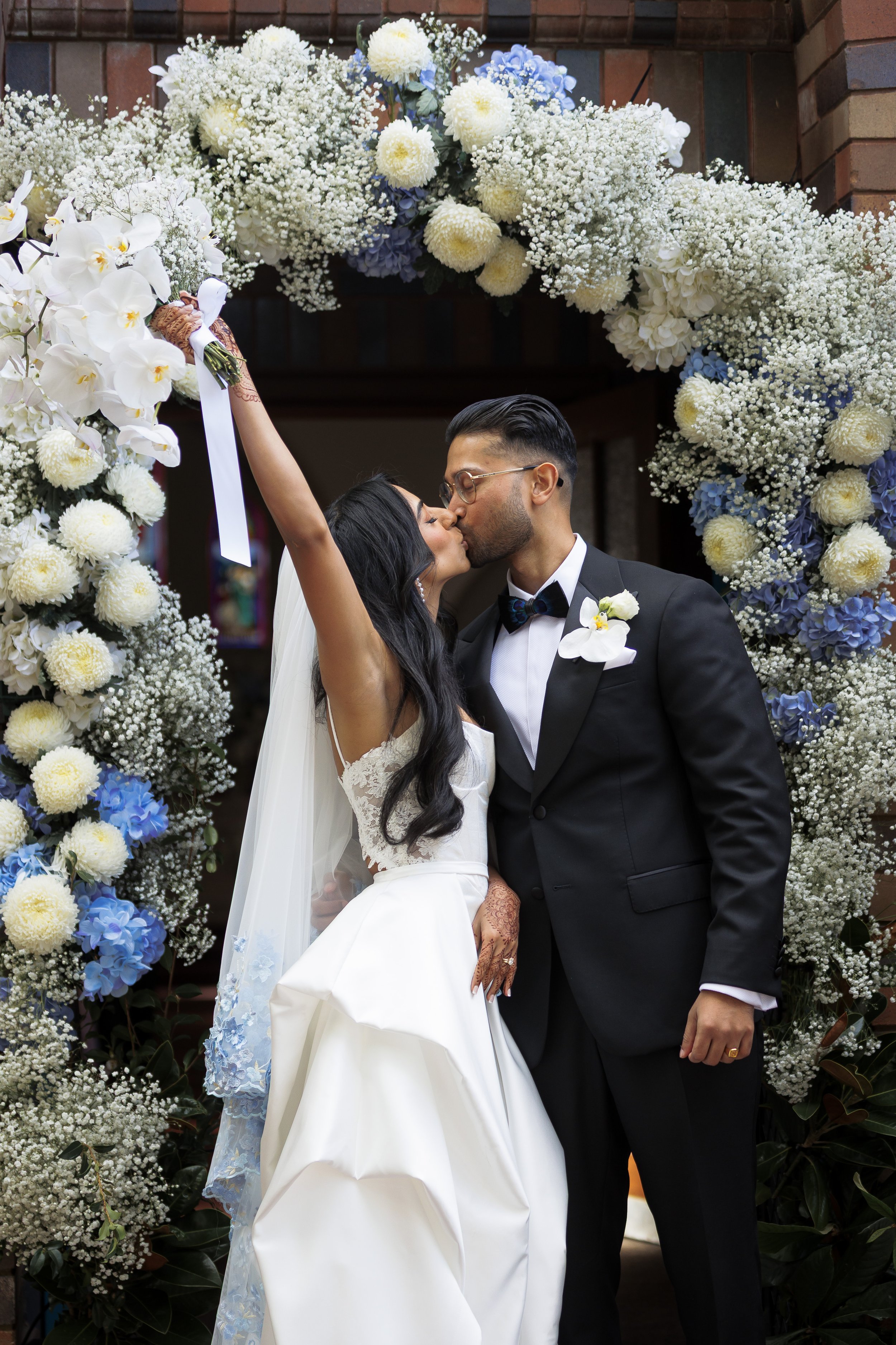 Bride and groom sharing a kiss in front of a floral wedding arch with white and blue flowers, celebrating their marriage.