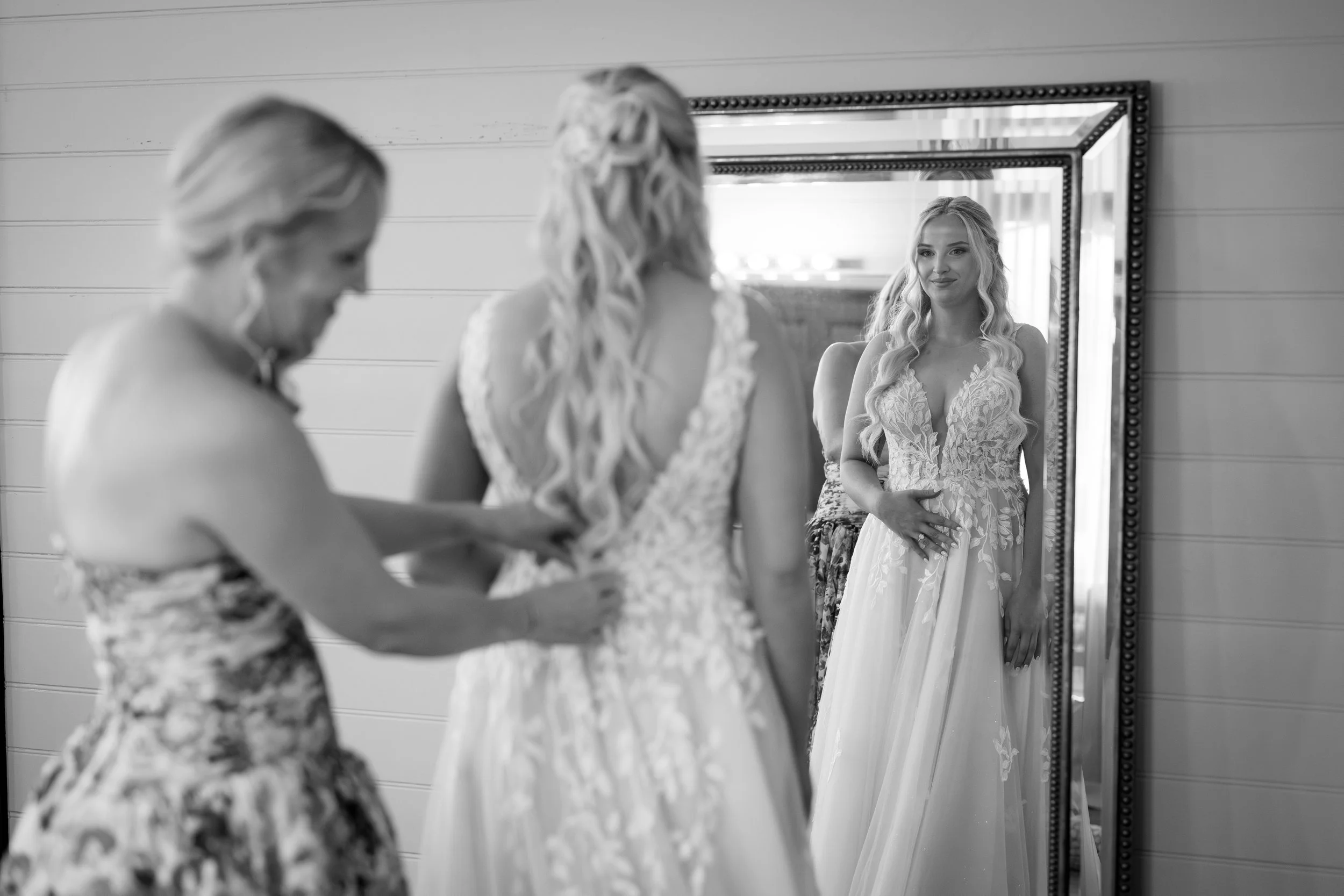 A woman in a wedding dress looking at her reflection in a mirror, while another woman helps her with the dress.