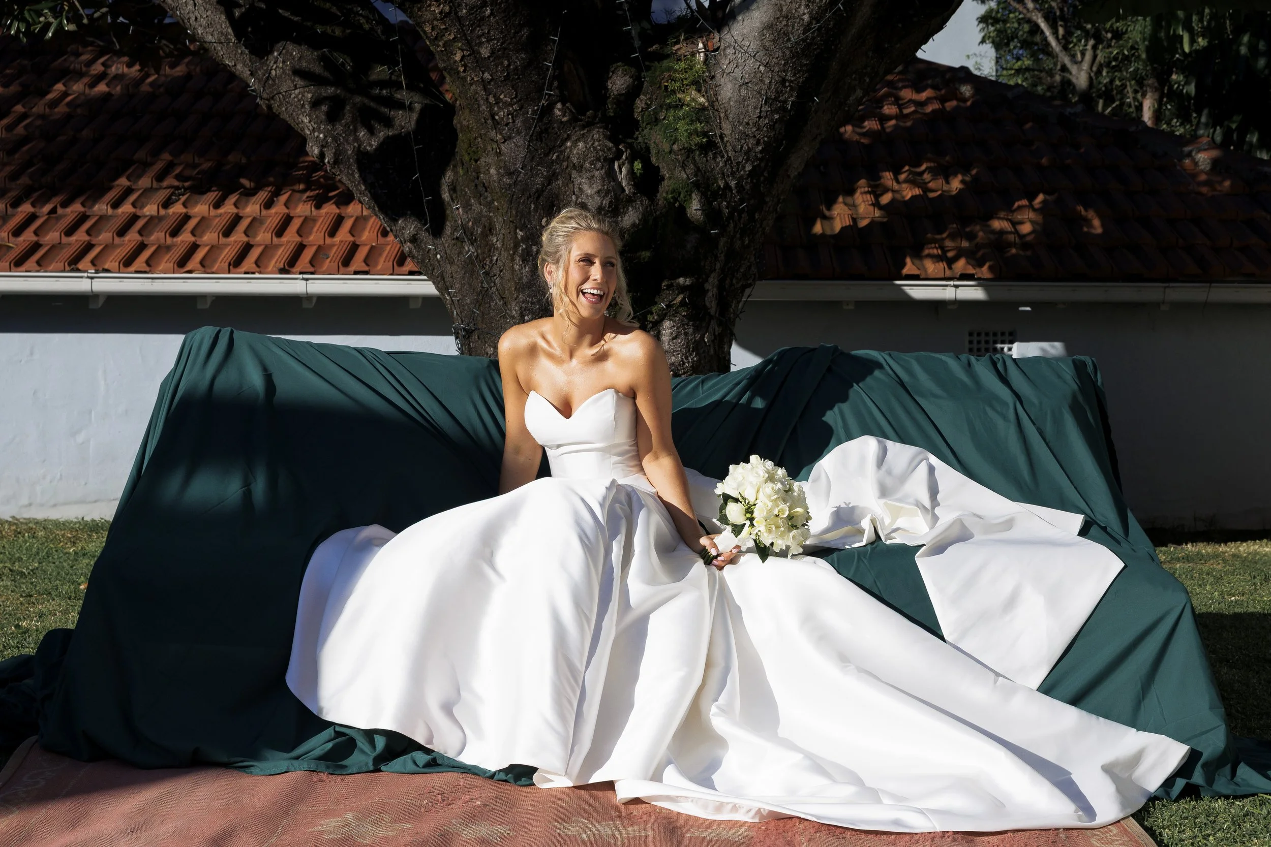 A bride in a white wedding gown sitting on a green-covered outdoor sofa, holding a bouquet of white flowers, smiling, with a large tree and a building with a red-tiled roof in the background on a sunny day.