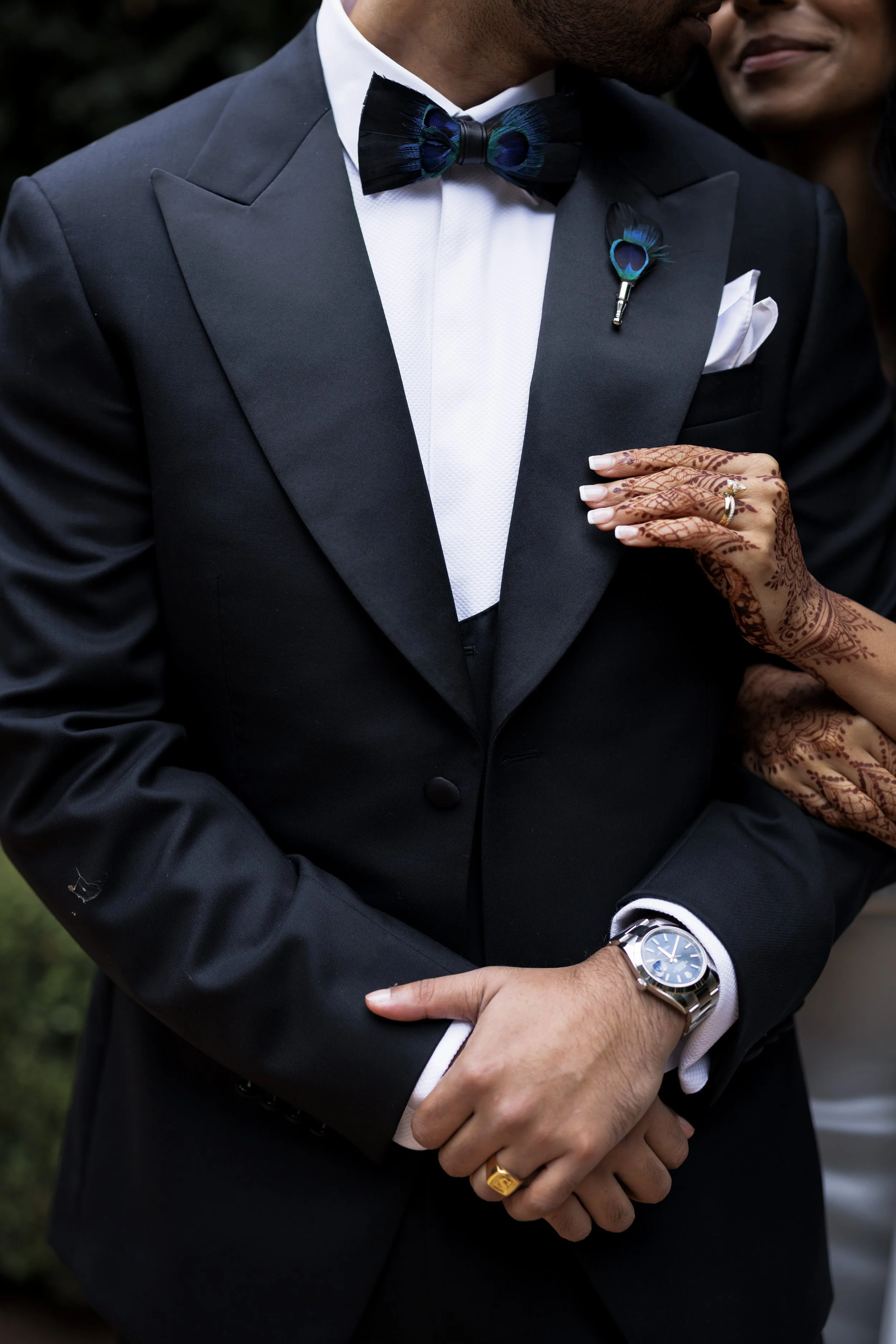 Close-up of a man in a black tuxedo with a white shirt and black bow tie, with a peacock feather boutonniere. He's wearing a silver watch, gold ring, and has henna designs on his hand, with a woman adjusting his sleeve.