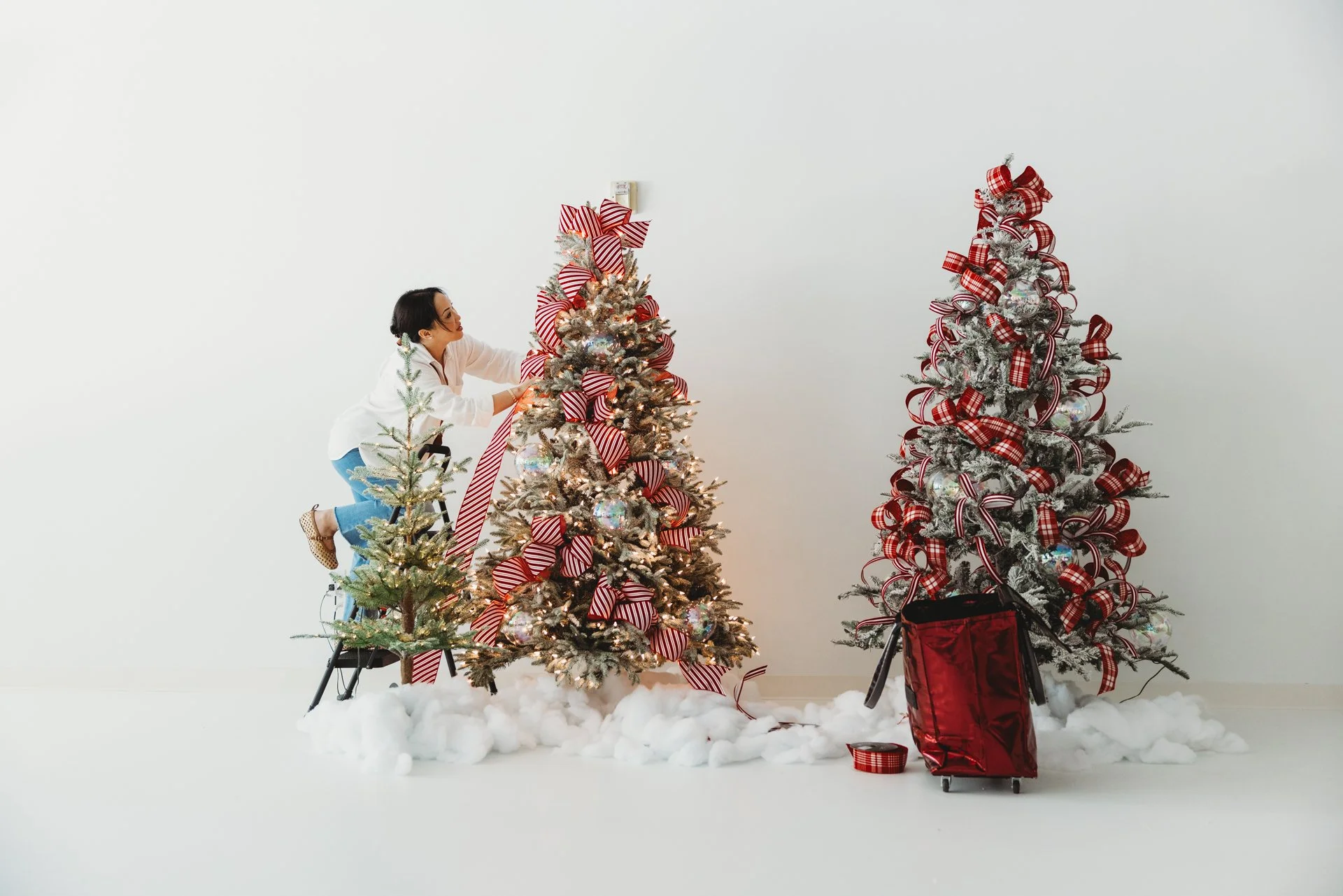 Woman decorating a Christmas tree with red and white ribbons in a blank room with white background.