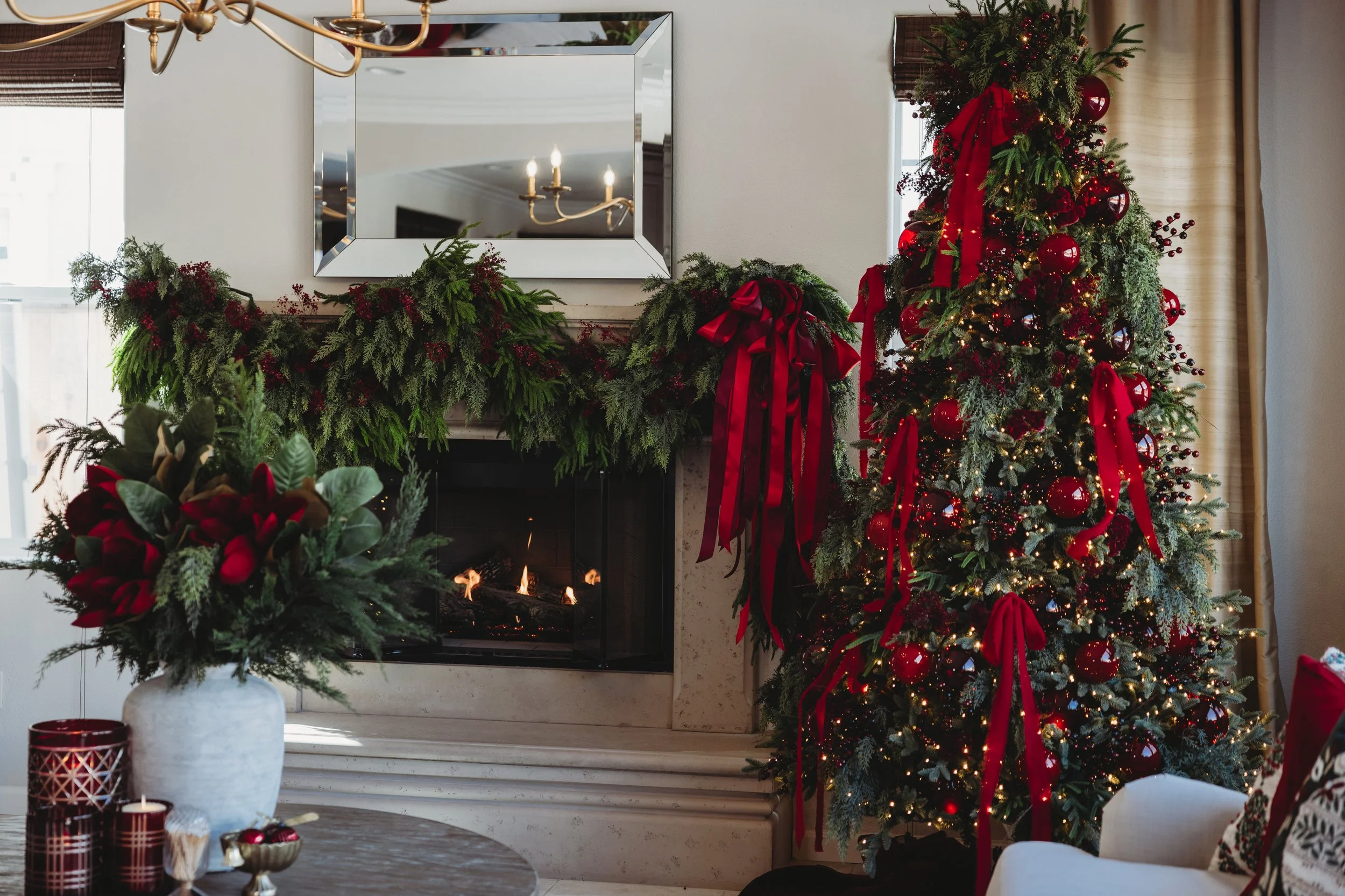 A decorated living room with a Christmas tree adorned with red ornaments, ribbons, and lights. A fireplace with a mirror above it and festive greenery garland, candles, and flower arrangements in the foreground.
