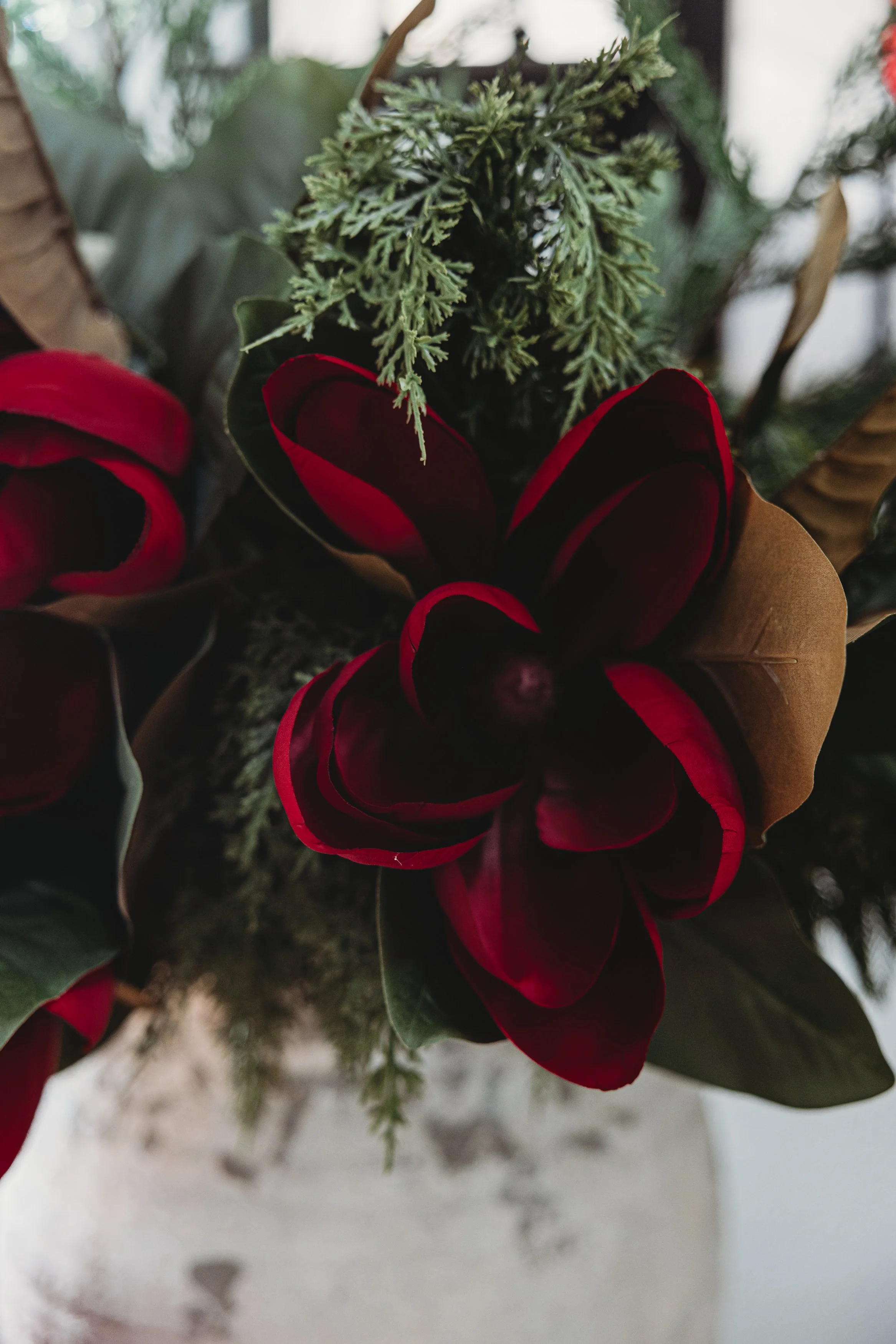 Close-up of a floral arrangement featuring red and dark red petals, green coniferous foliage, and other greenery in a rustic vase.