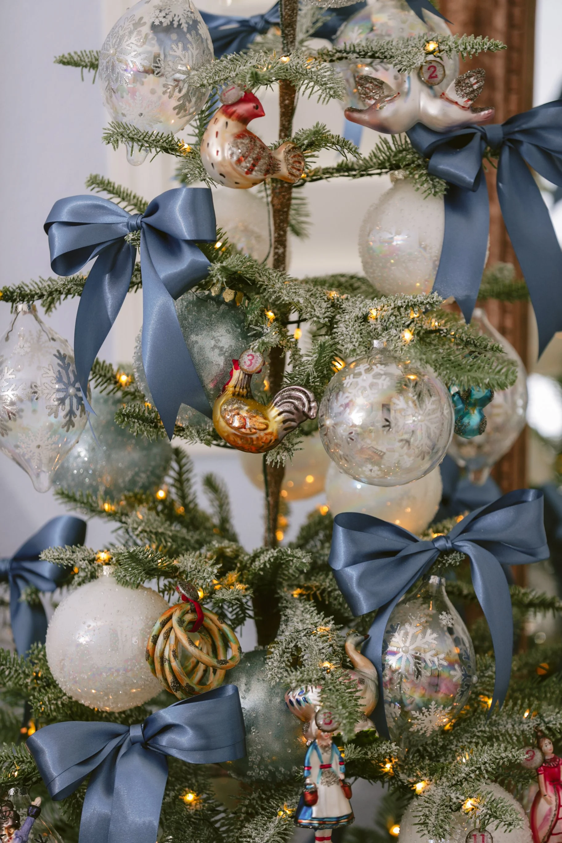 Decorated Christmas tree with ornaments including glass balls, ceramic birds, ribbons, and figurines, illuminated with string lights.