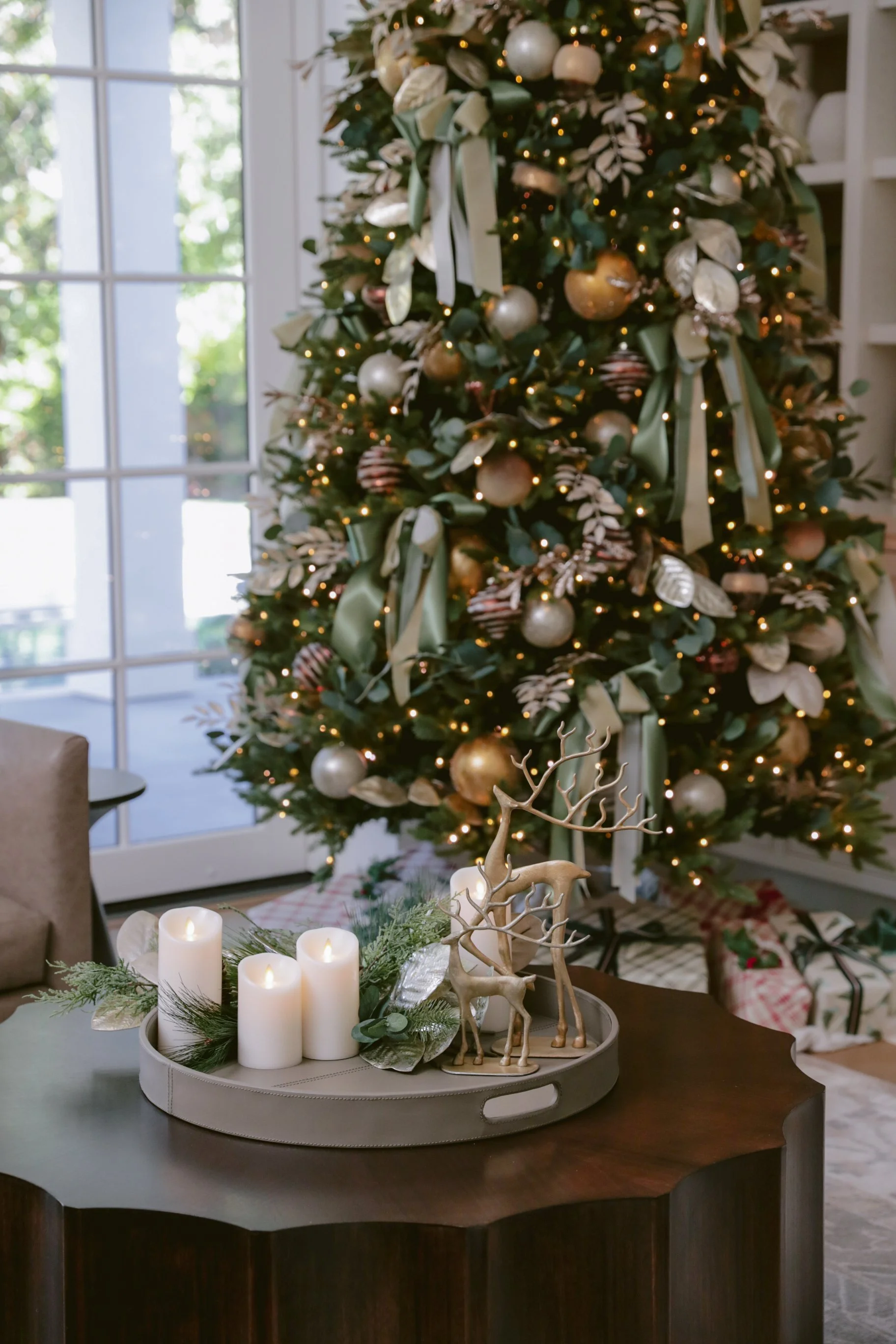 Decorated Christmas tree with gold and green ornaments, ribbons, and lights in a cozy room with large windows.