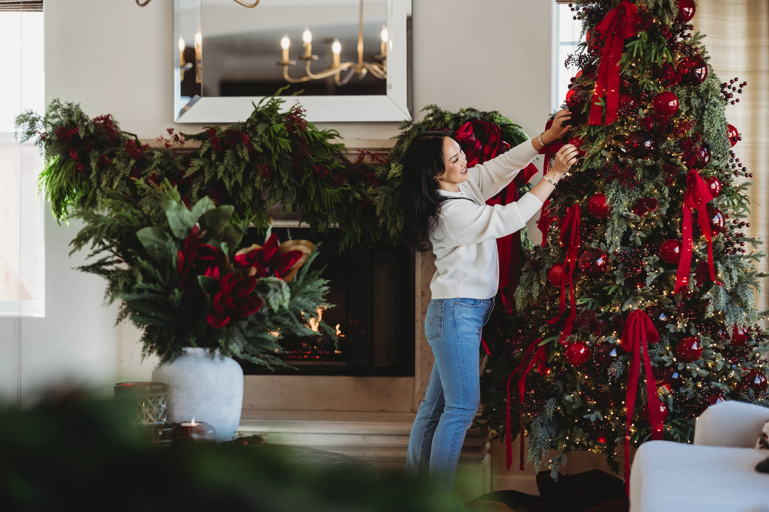 A woman decorating a Christmas tree with red ornaments and ribbons in a cozy, well-lit living room with holiday decorations.
