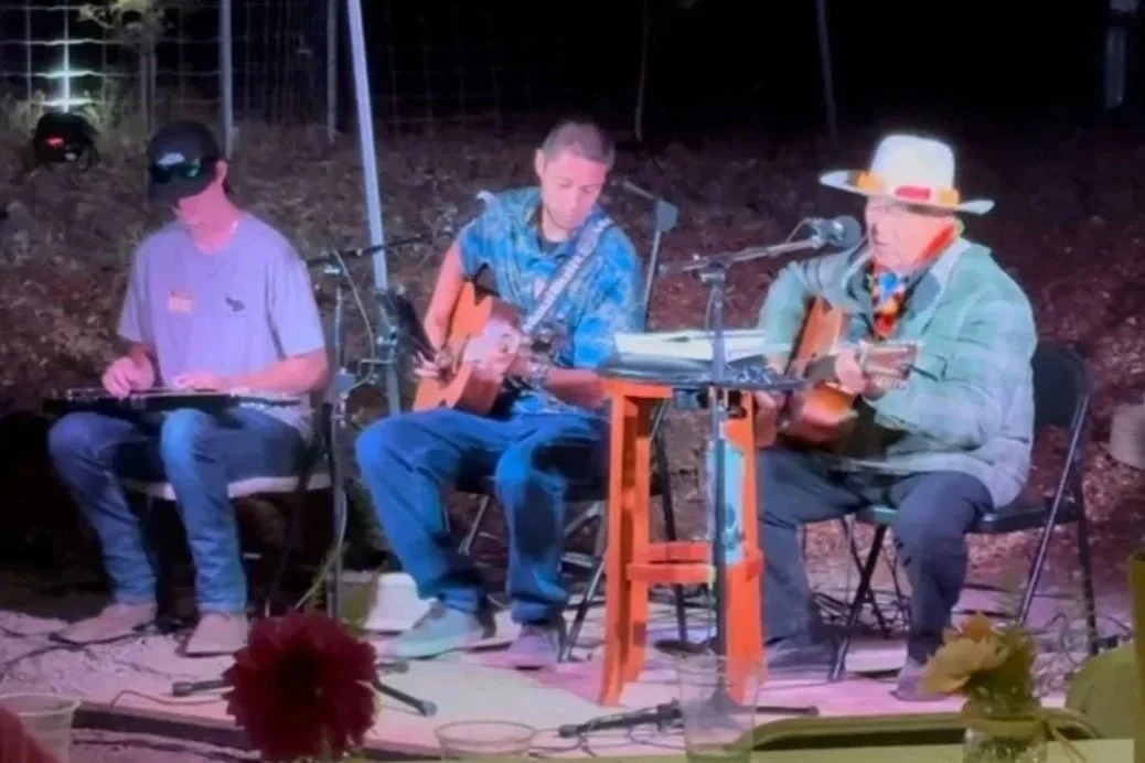 Three men sitting outdoors at night, playing guitars and a small percussion instrument, with microphones and music sheets, under string lights.