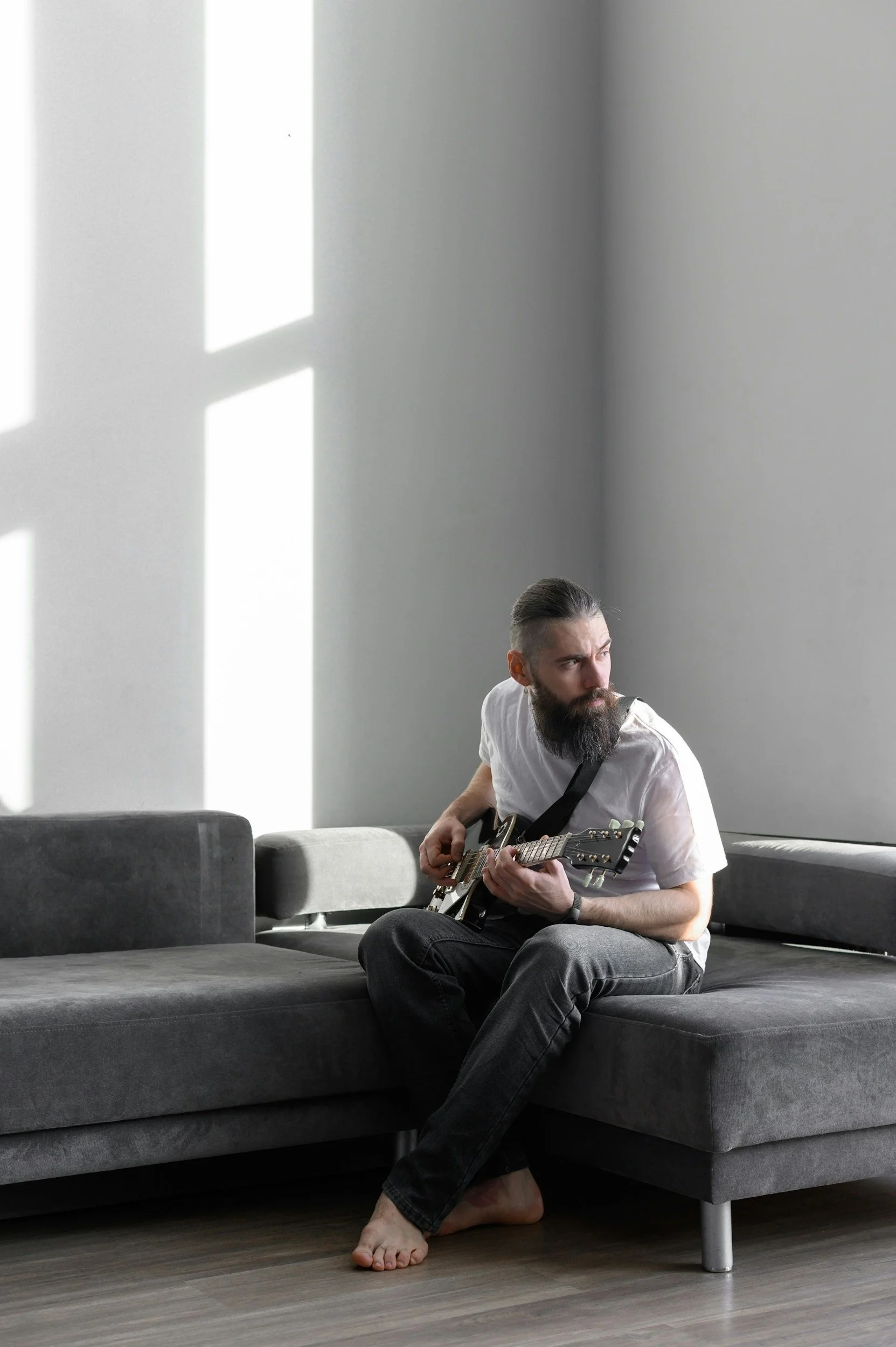 A man with a beard and long hair sitting on a gray sectional sofa, playing an electric guitar, with sunlight streaming through windows and casting shadows on the wall.