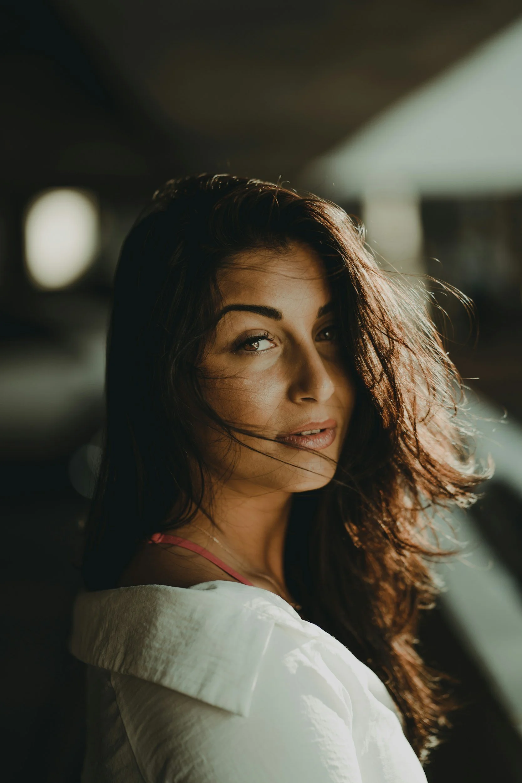 A woman with wavy brown hair and light skin looking over her shoulder in a parking garage, sunlight illuminating her face.