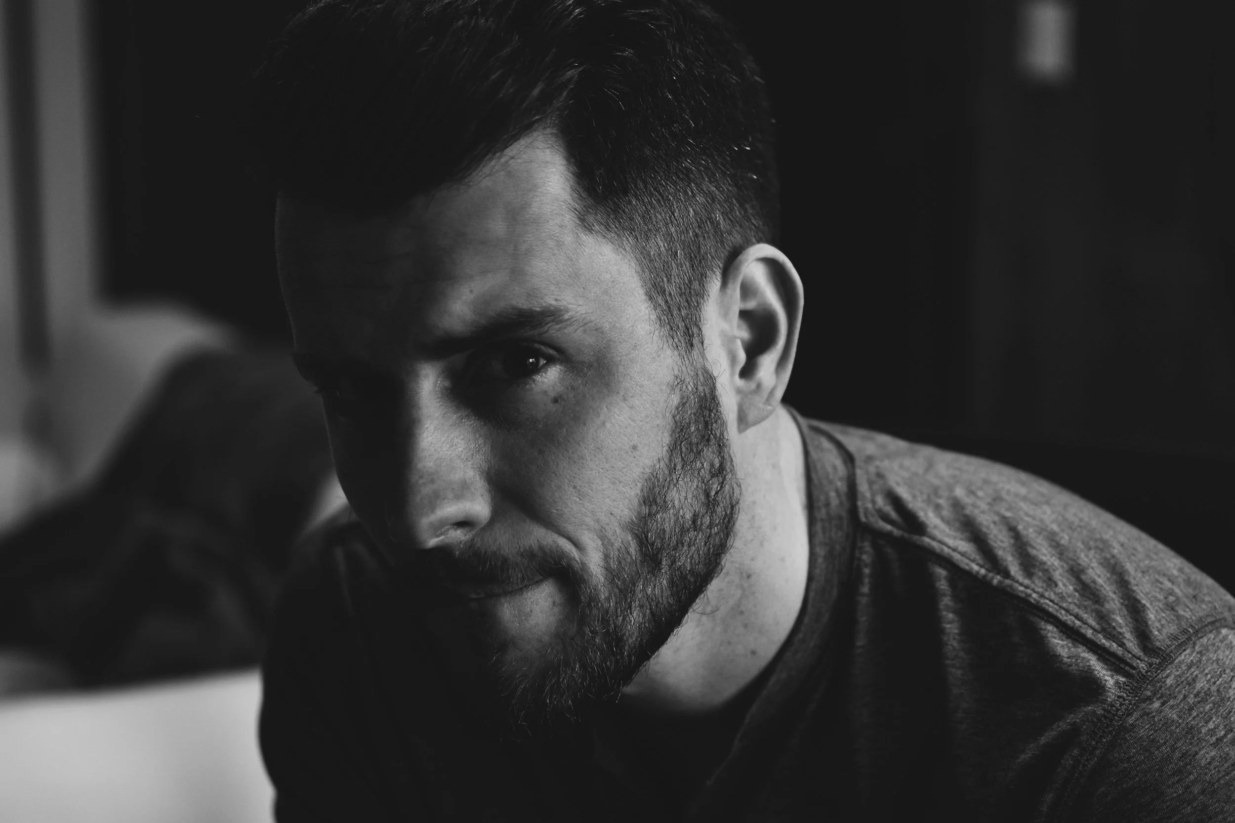 Black and white close-up photograph of a man with short hair and a beard, looking at the camera with a serious expression.