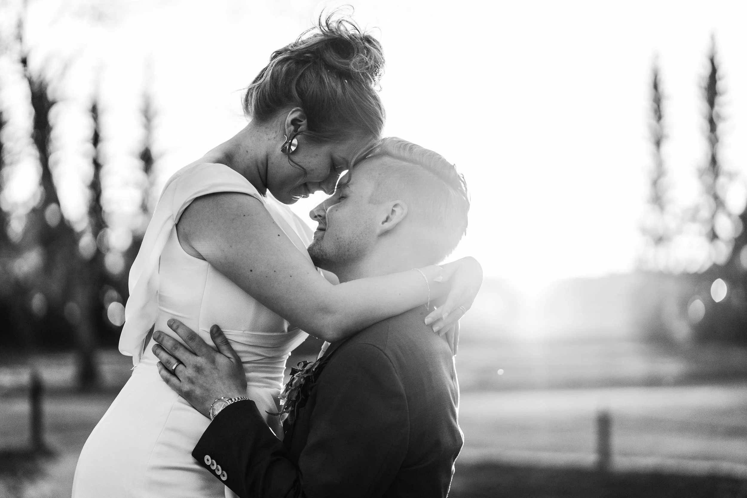 A black-and-white photo of a couple sharing a romantic moment outdoors, with the woman standing and the man kneeling, their foreheads touching, in a setting with trees and sunlight in the background.