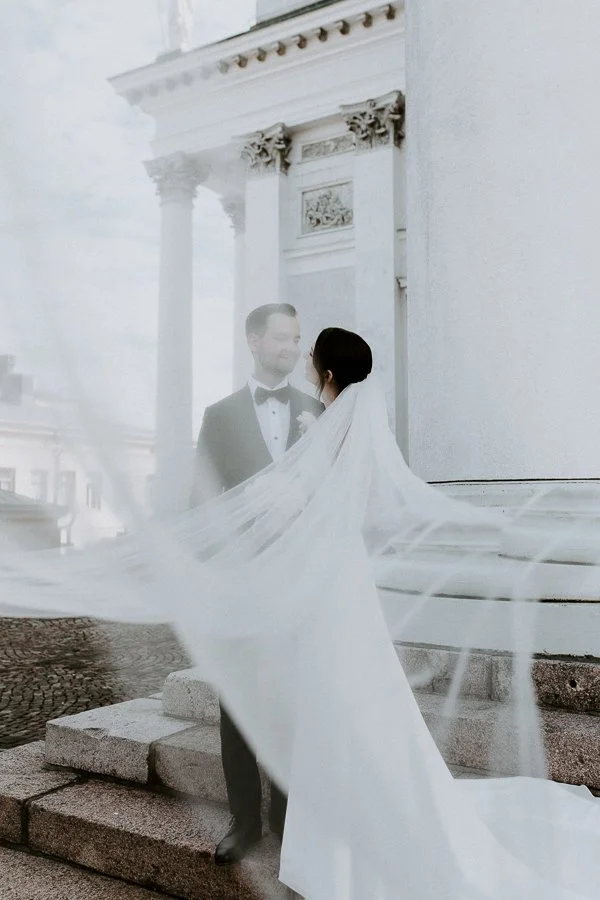 A bride and groom standing outside a historic building with columns, sharing a moment while the bride's veil is flowing in the wind.