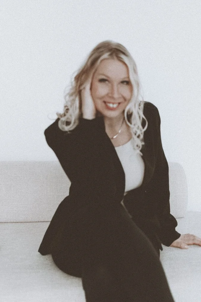 A woman with blonde curly hair wearing a black blazer and white shirt, smiling and sitting on a white surface against a plain white background.