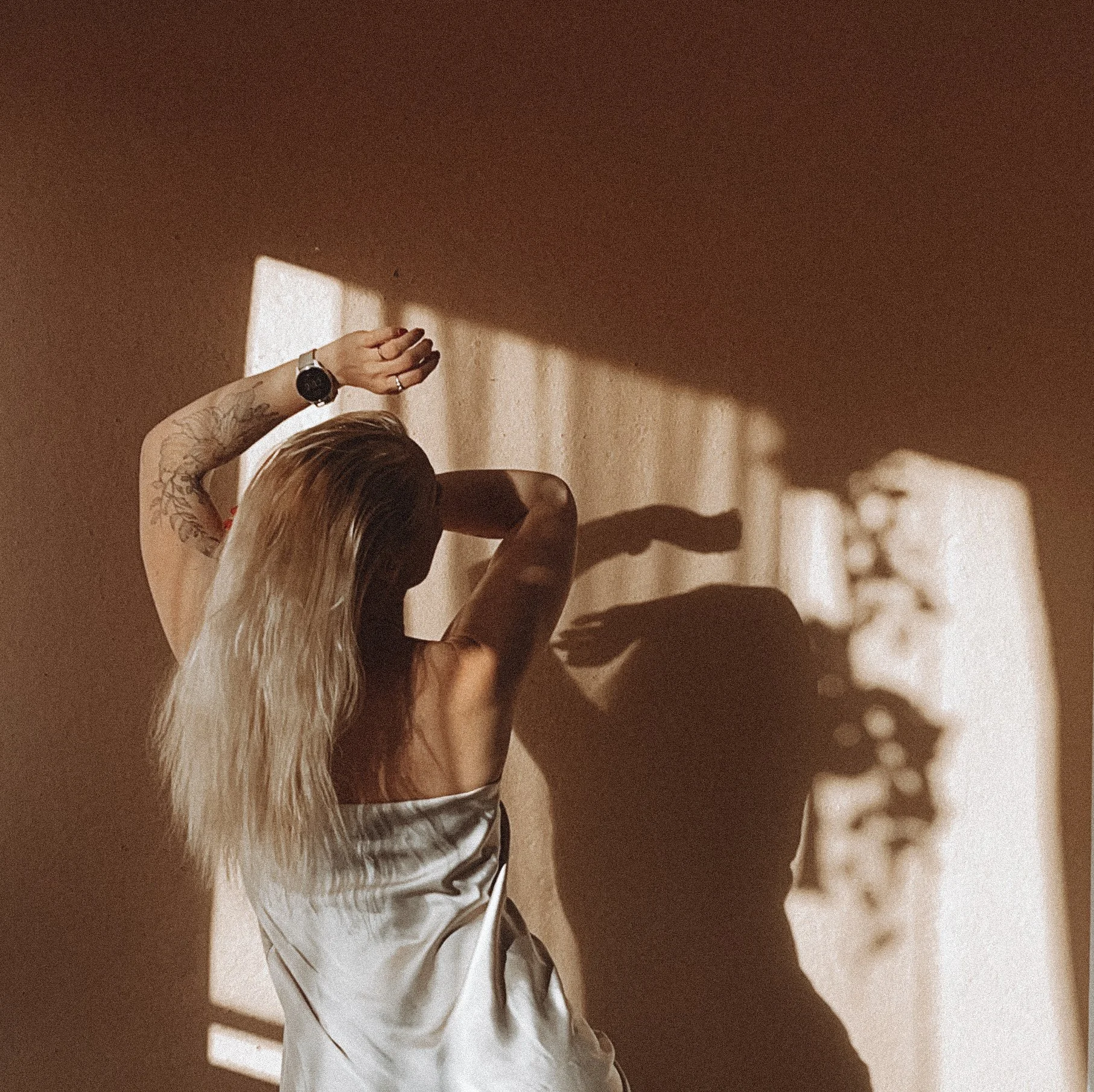A woman with long blonde hair, tattoos on her arm, wearing a silky dress, standing in front of a wall with shadows and sunlight coming through a barred window.