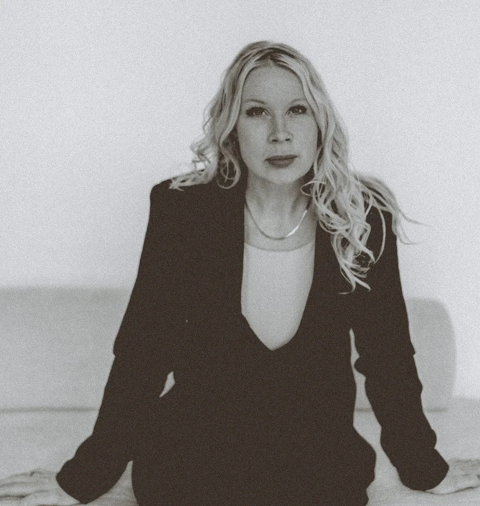 A woman with long, wavy blonde hair, wearing a black blazer and a light-colored top, sitting on a white surface against a plain background.