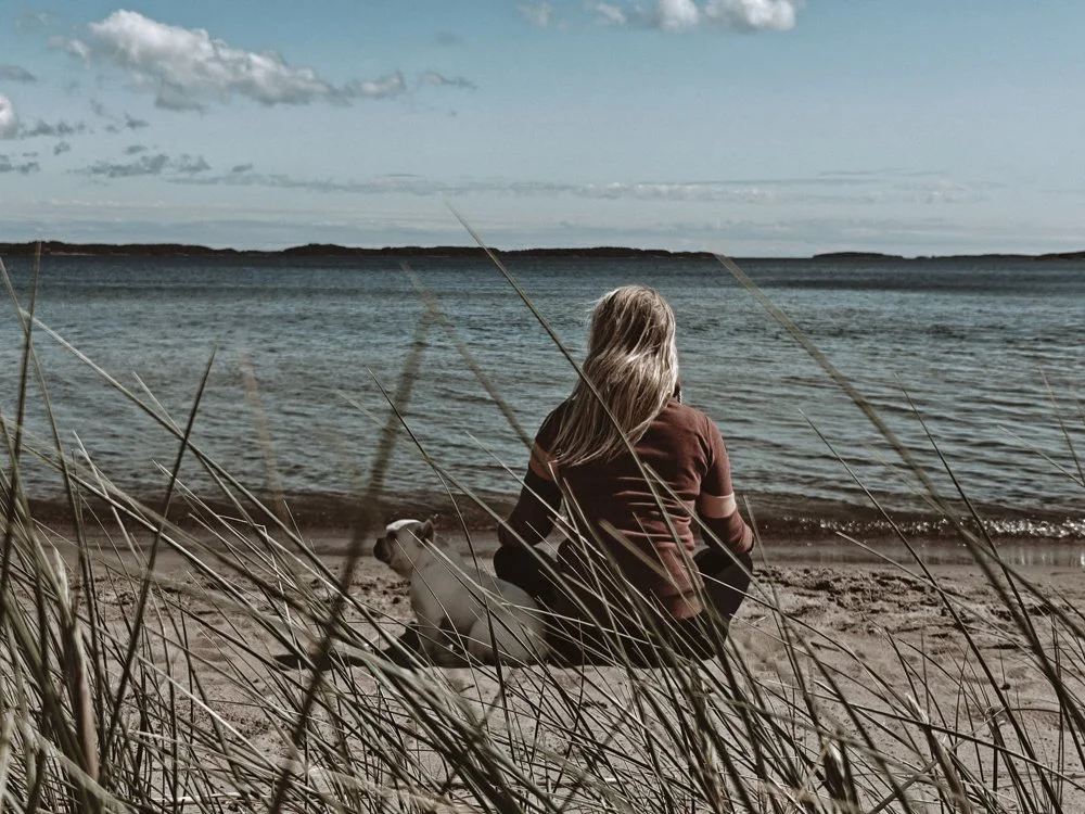 A person with blonde hair sitting on the sandy beach next to a dog, looking out at the water on a partly cloudy day.