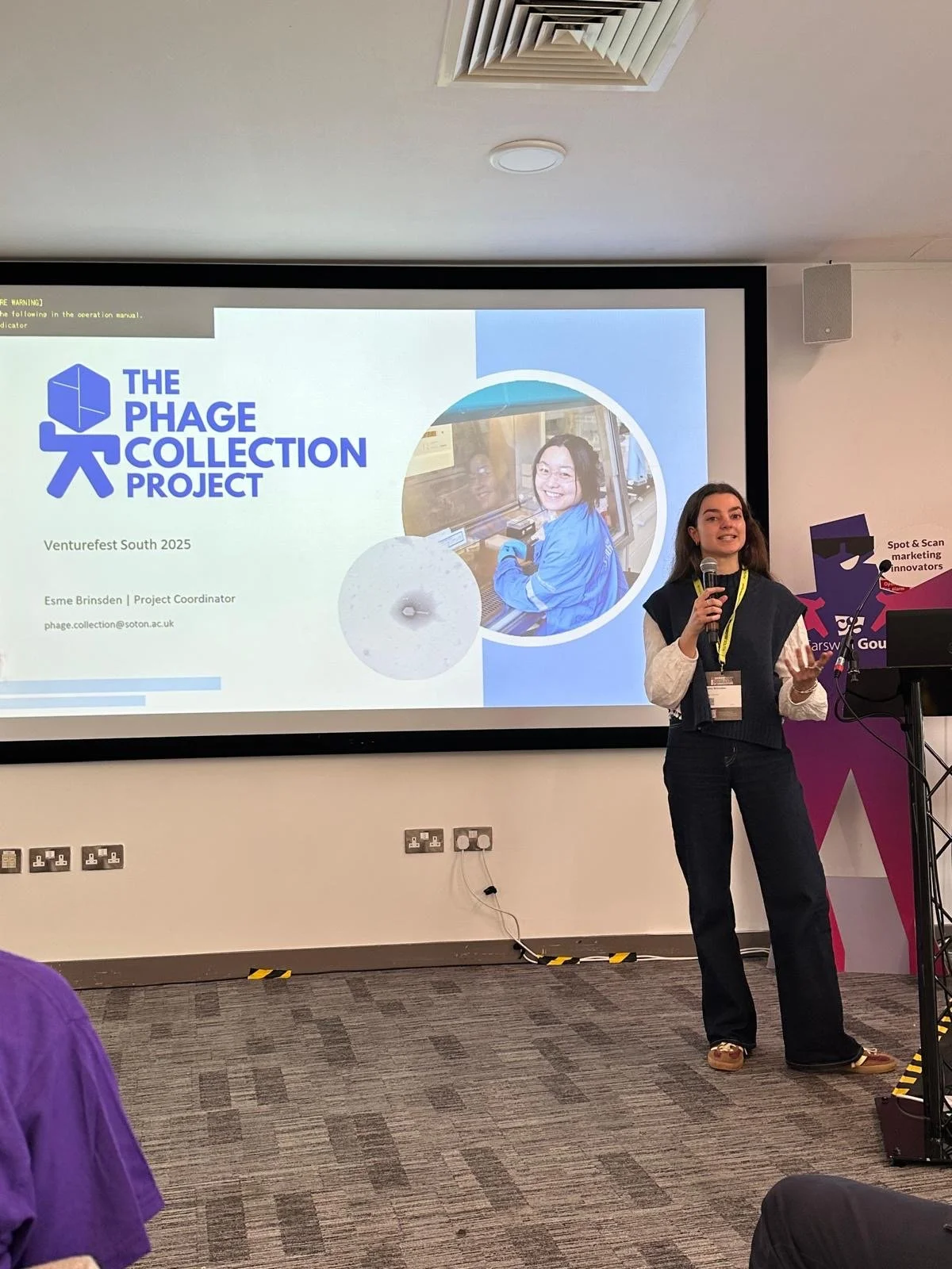 A woman is giving a presentation at Venturefest South 2023, standing near a large screen displaying a slide about 'The Phage Collection Project' with a photo of a smiling woman in a blue lab coat working with laboratory equipment.