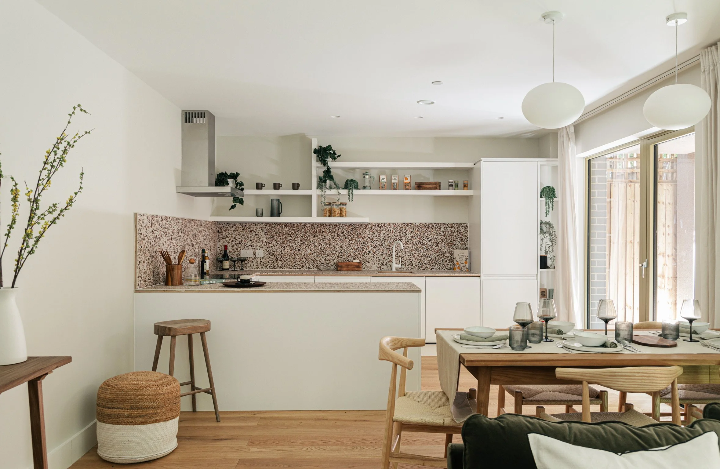 Open-plan kitchen and dining area with white cabinetry, open shelves, and a mosaic backsplash, decorated with plants, a dining table set with plates and glasses, near a large window with curtains.