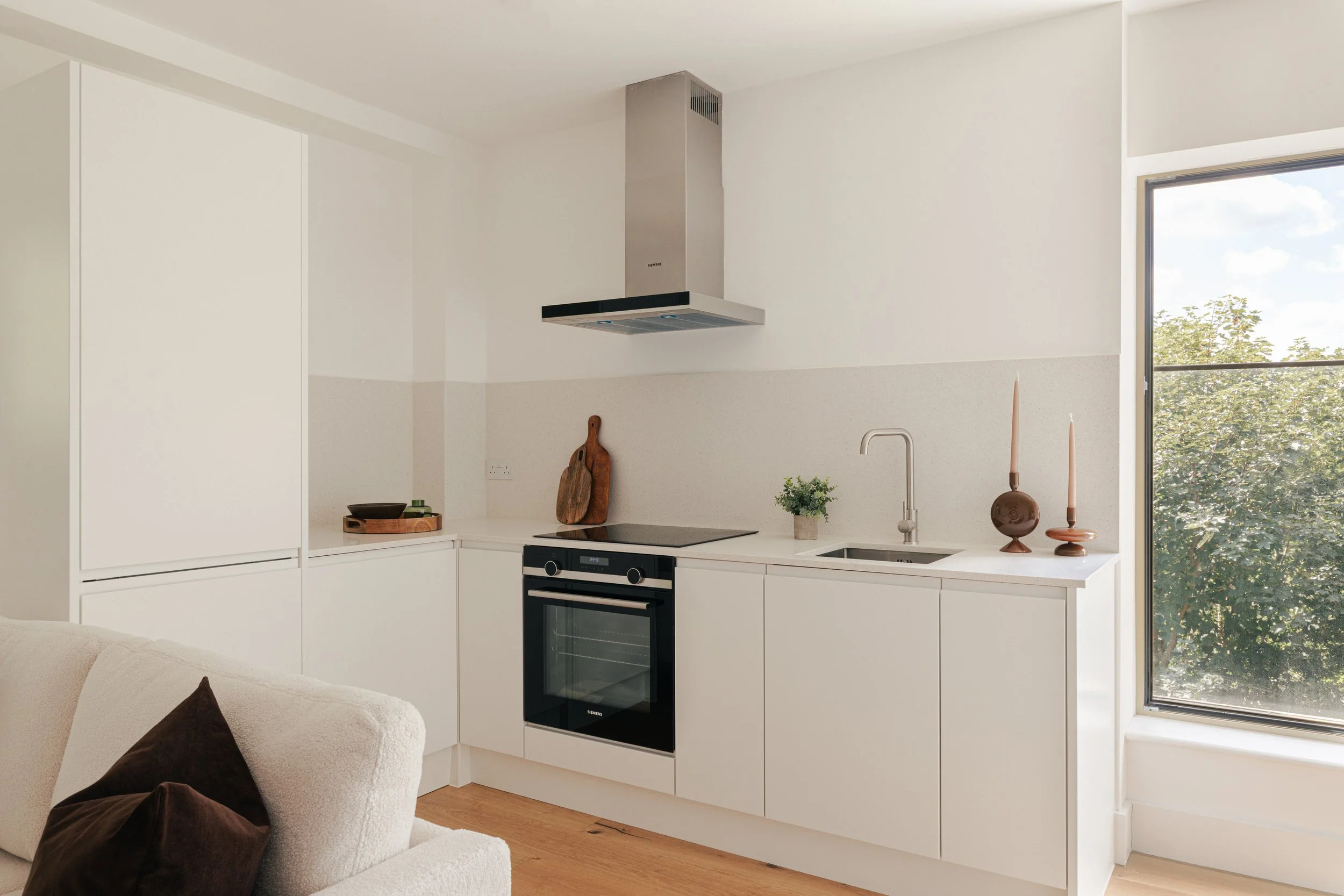 Minimalist white kitchen with built-in oven, black stovetop, stainless steel range hood, small potted plant, wooden cutting boards, and pink candles near large window.