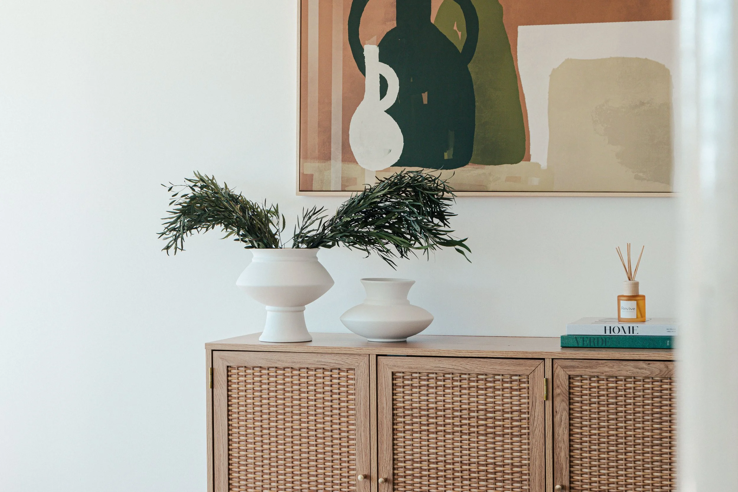 A wooden cabinet with woven rattan doors, decorated with two white vases containing green branches, a small amber reed diffuser, and stacked books on a white wall with a framed abstract art piece.