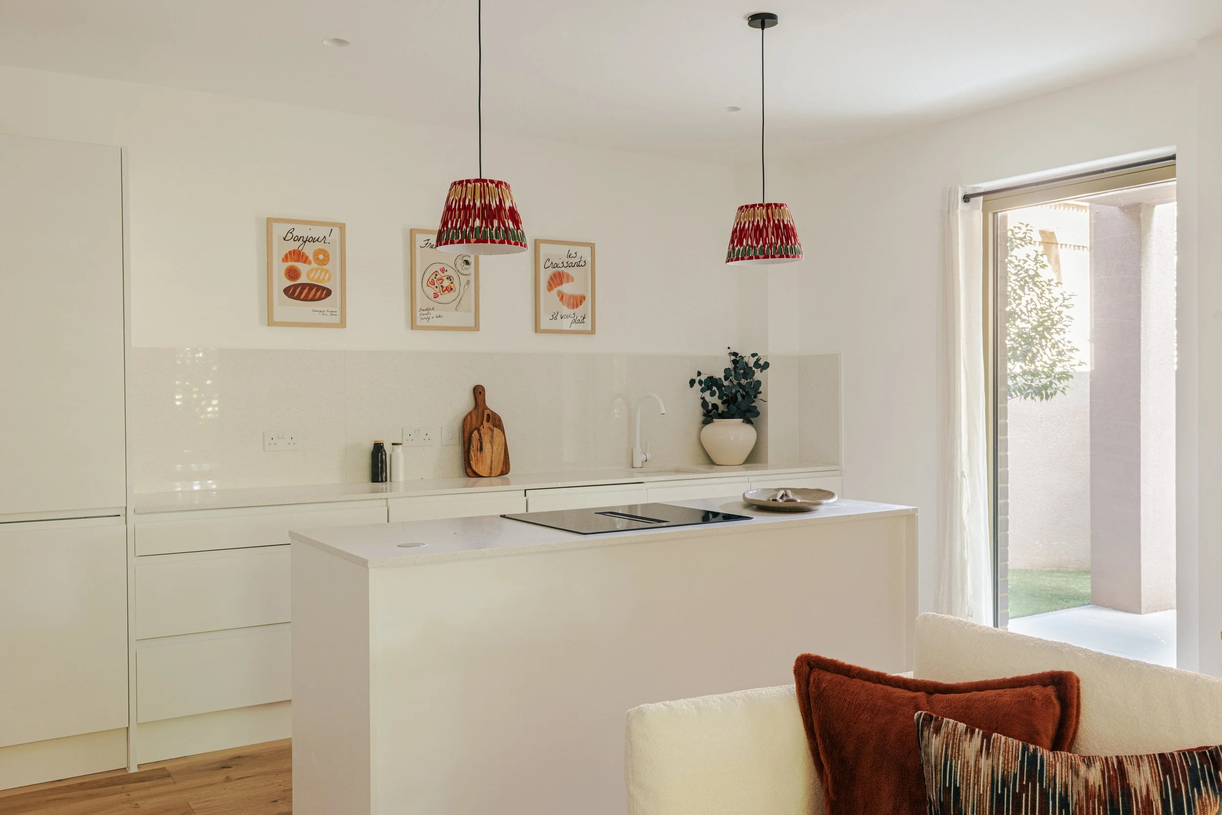 Modern kitchen with white cabinetry, a white kitchen island, and a sliding glass door leading outside, decorated with three framed art prints and two colorful hanging lamps.