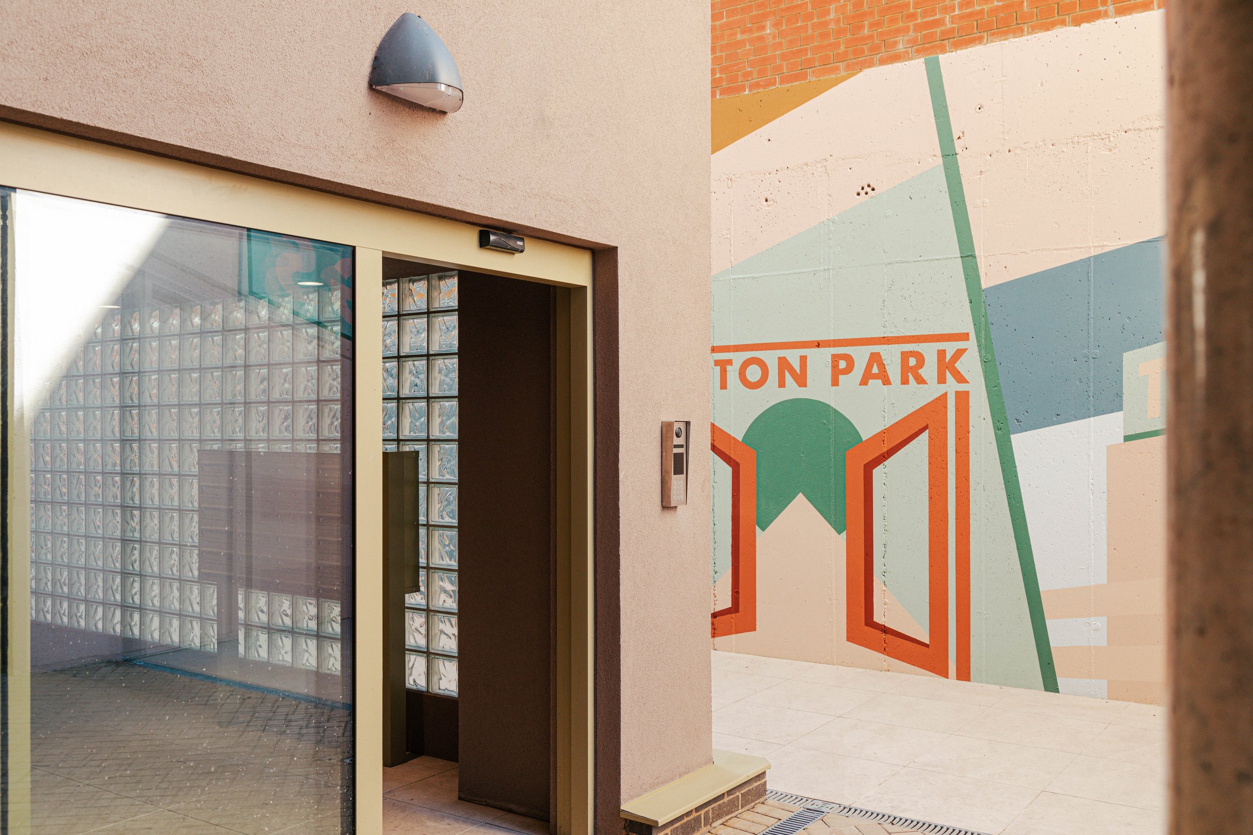 Entrance to a building with glass block wall, elevator with beige frame, and a mural on the exterior wall with green, orange, and beige geometric shapes and the text "TUN PARK".