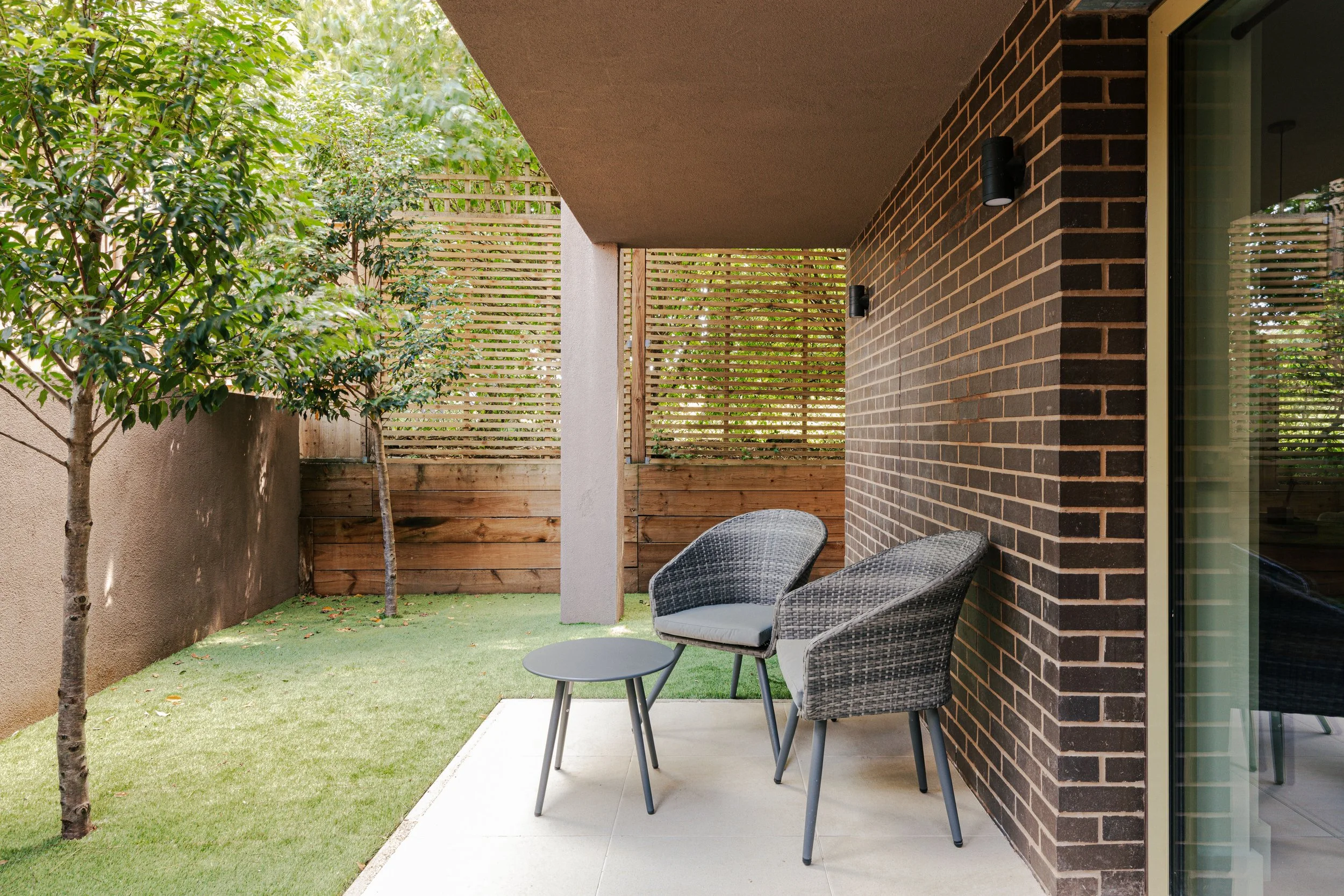 Outdoor patio with two gray wicker chairs and a small gray table, green grass, trees, and a wooden privacy fence.