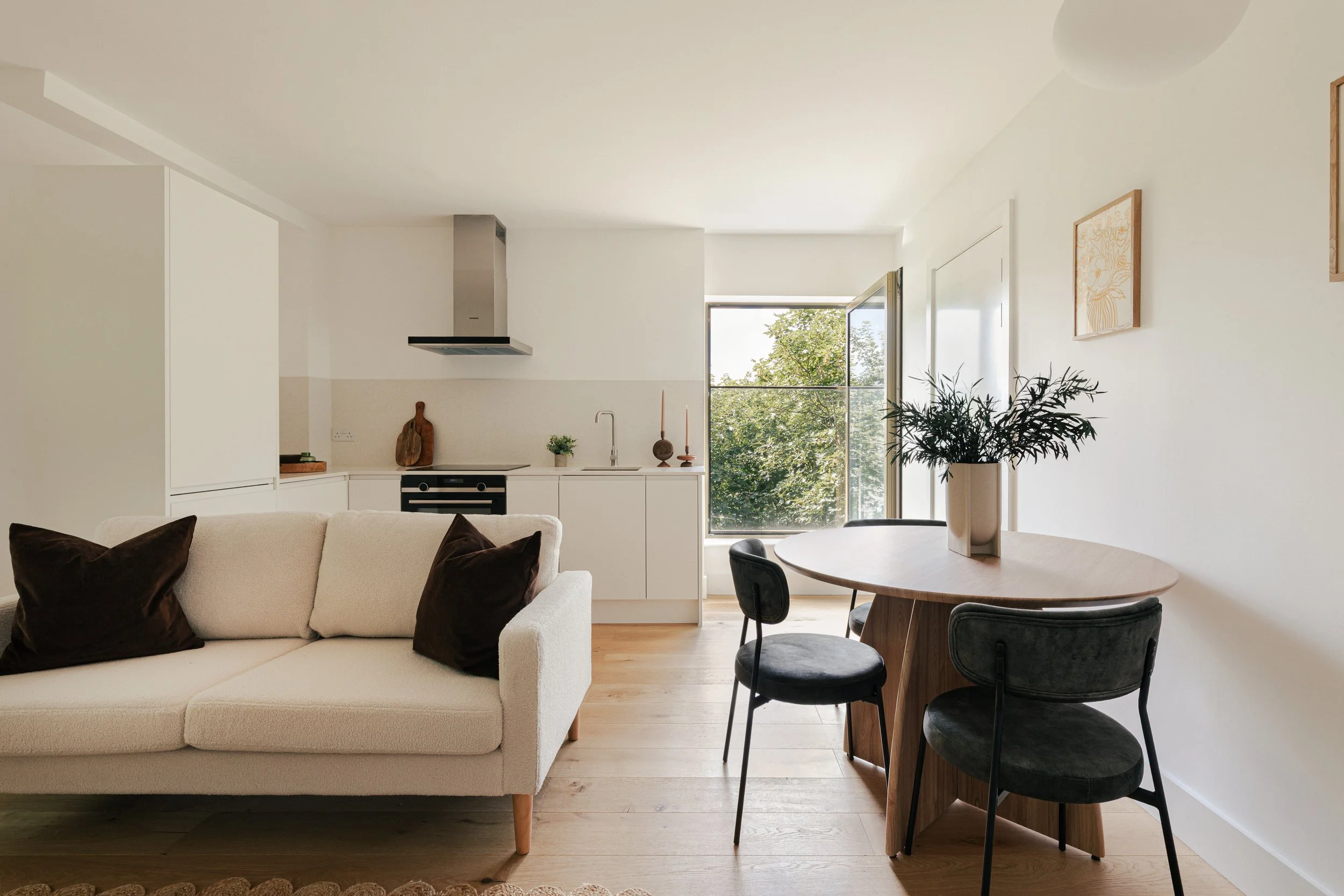 View of a modern, minimalistic open-plan living room and kitchen with a white sofa, black cushions, a round wooden dining table with black chairs, a potted plant, large window showing greenery outside, and white walls with simple artwork.