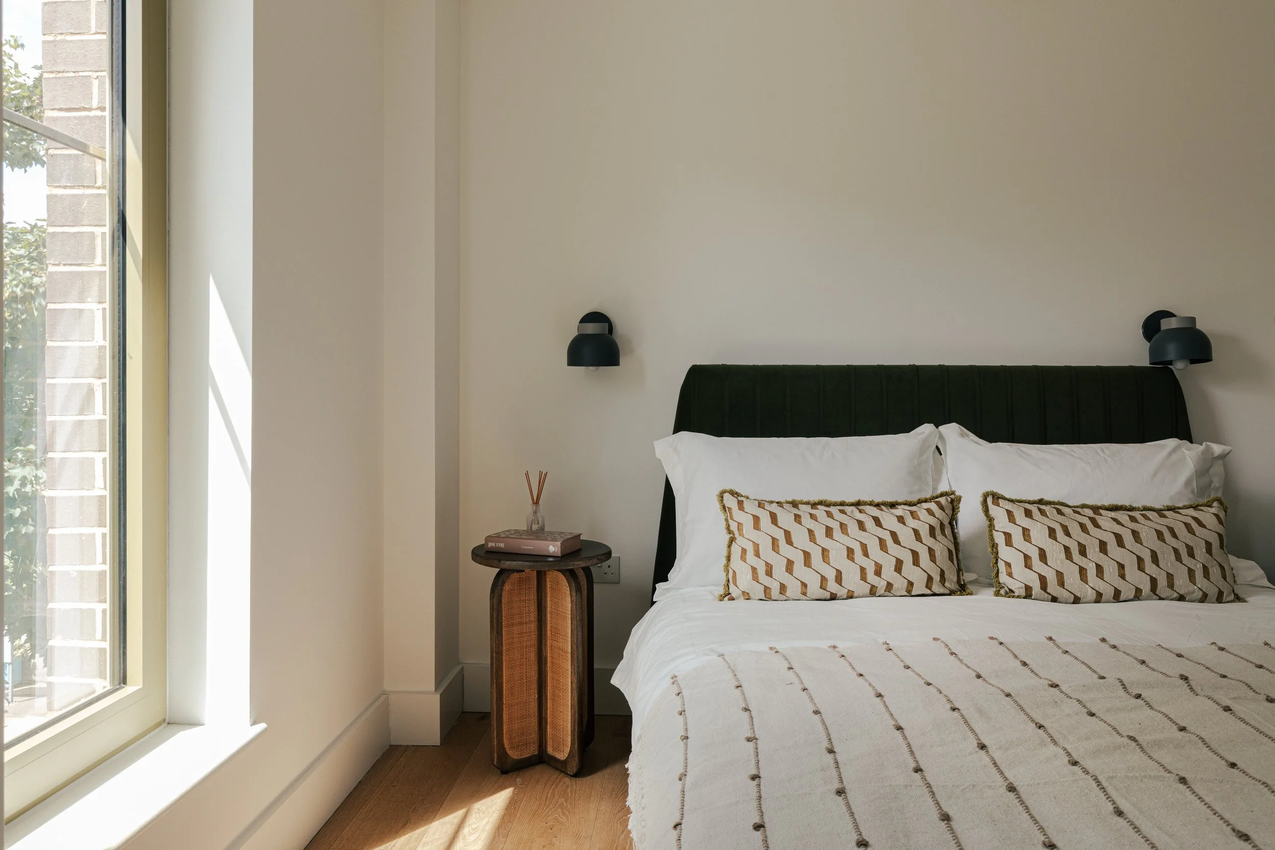 A neatly made bed with white bedding and two elongated decorative pillows with a geometric pattern, in a bedroom with a nightstand with reed diffuser and book, black wall-mounted lamps on a plain wall, and sunlight coming through a window.
