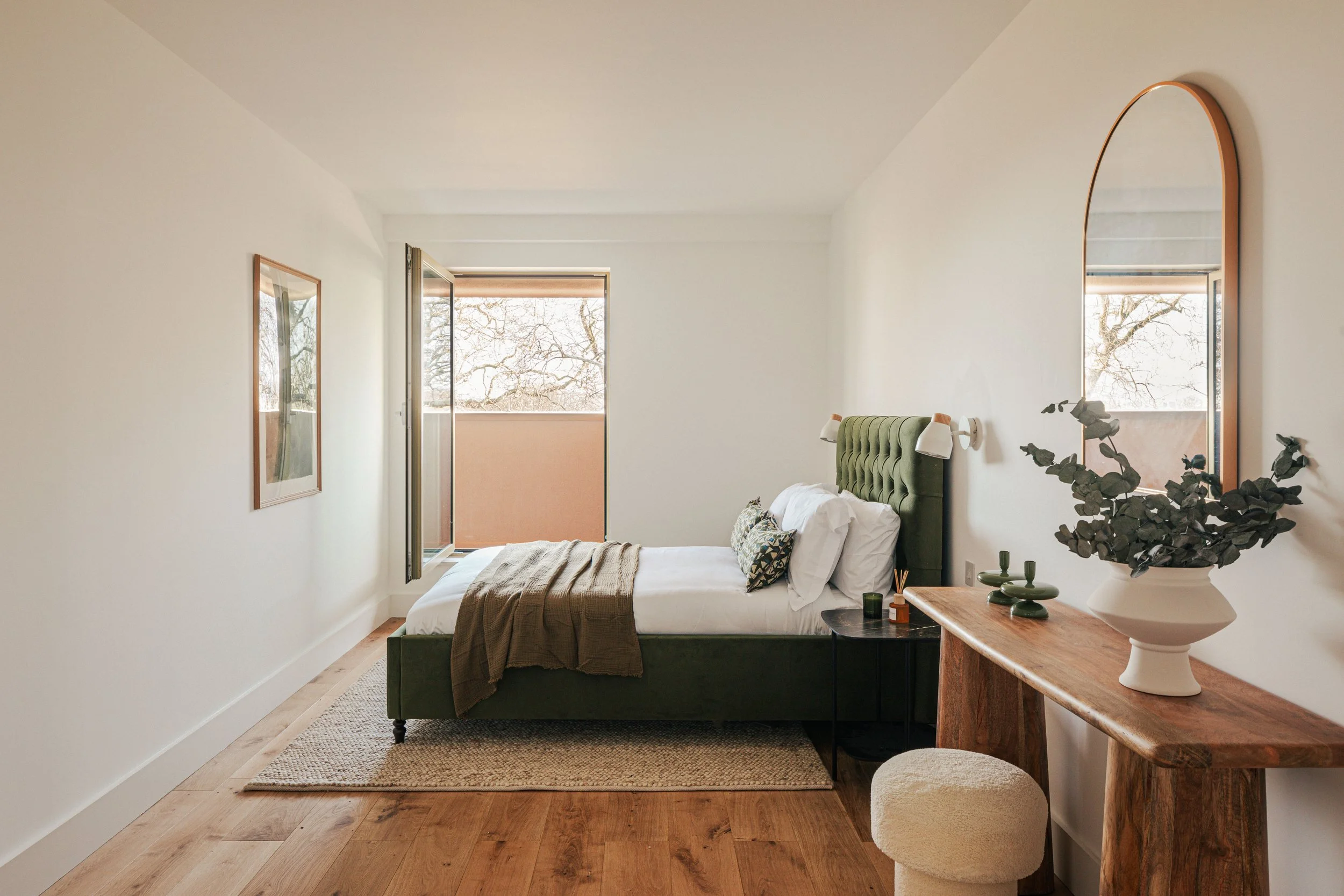 A bedroom with a green upholstered bed, a black side table, a wooden desk with a white vase and greenery, a mirror on the wall, a window with a balcony and tree branches outside, and a wooden floor.