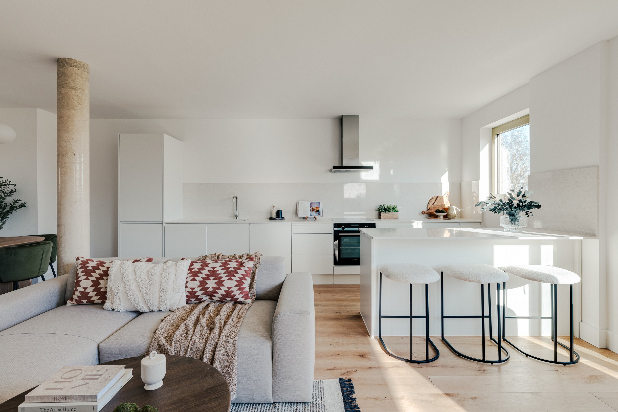 Modern, minimalist open-plan living room and kitchen in a bright home with white cabinets, a kitchen island with barstools, and a beige sofa with decorative pillows.