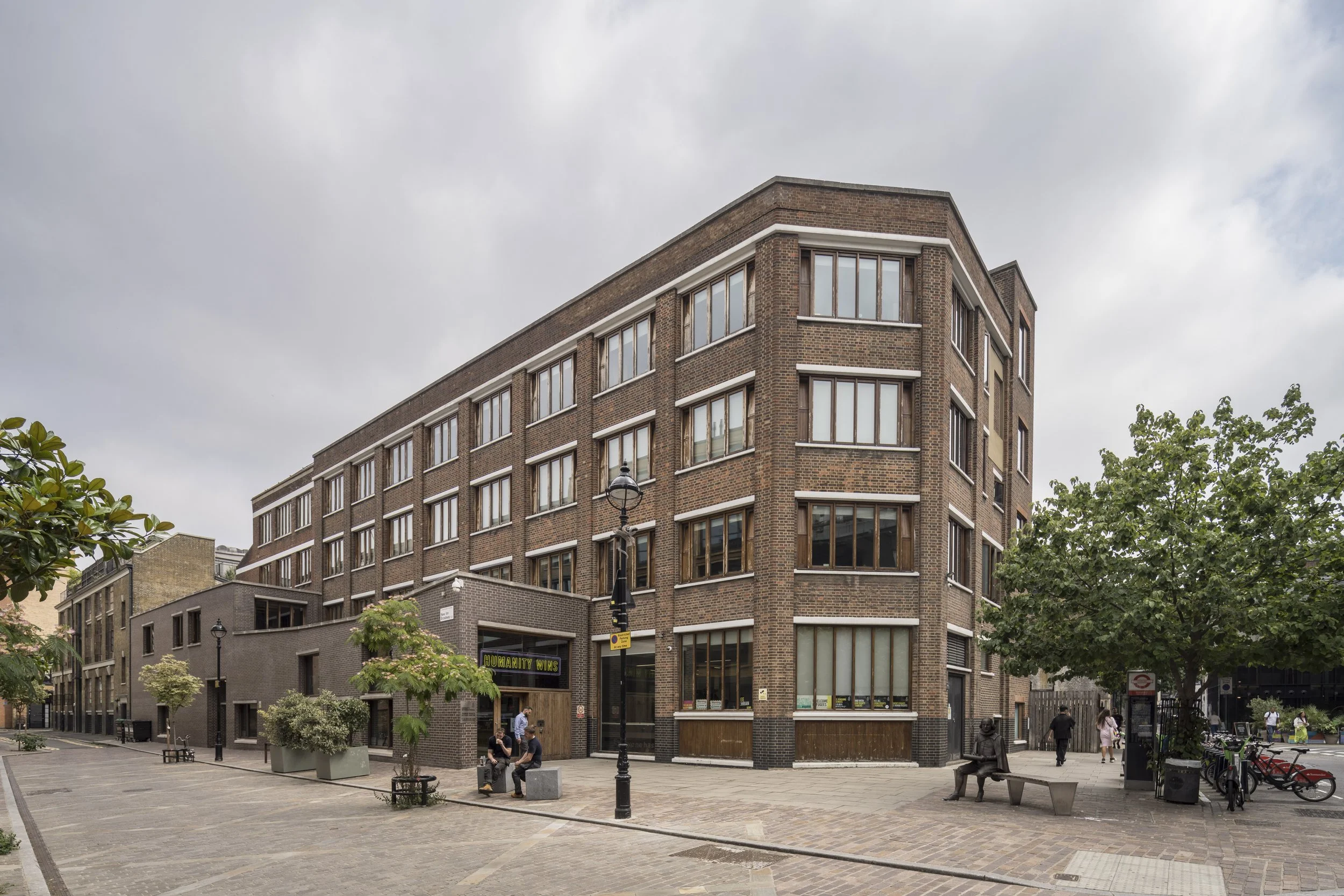 A multi-story brick building on a city street with a sidewalk, trees, benches, and people walking or sitting.