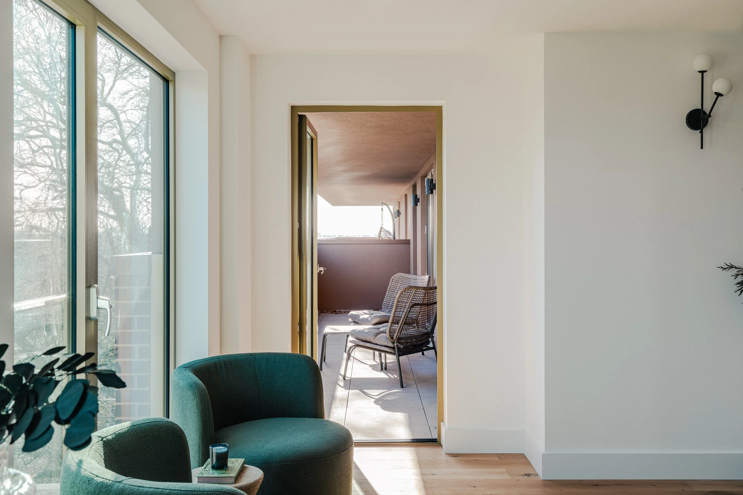 Interior view of a bright, modern living space with large glass sliding doors, a green armchair with a small table, and an outdoor balcony with chairs.
