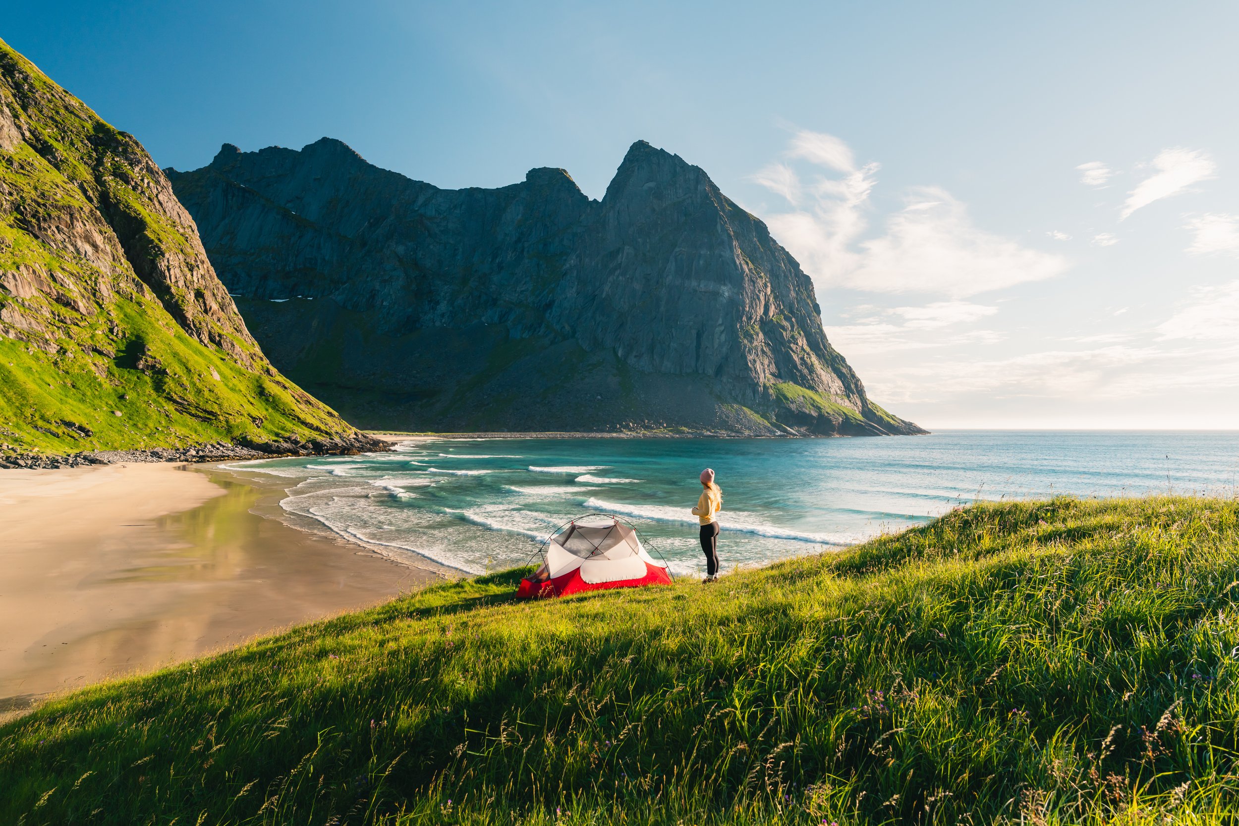 Person standing near a tent on a grassy hill overlooking a beach and ocean with mountains in the background.