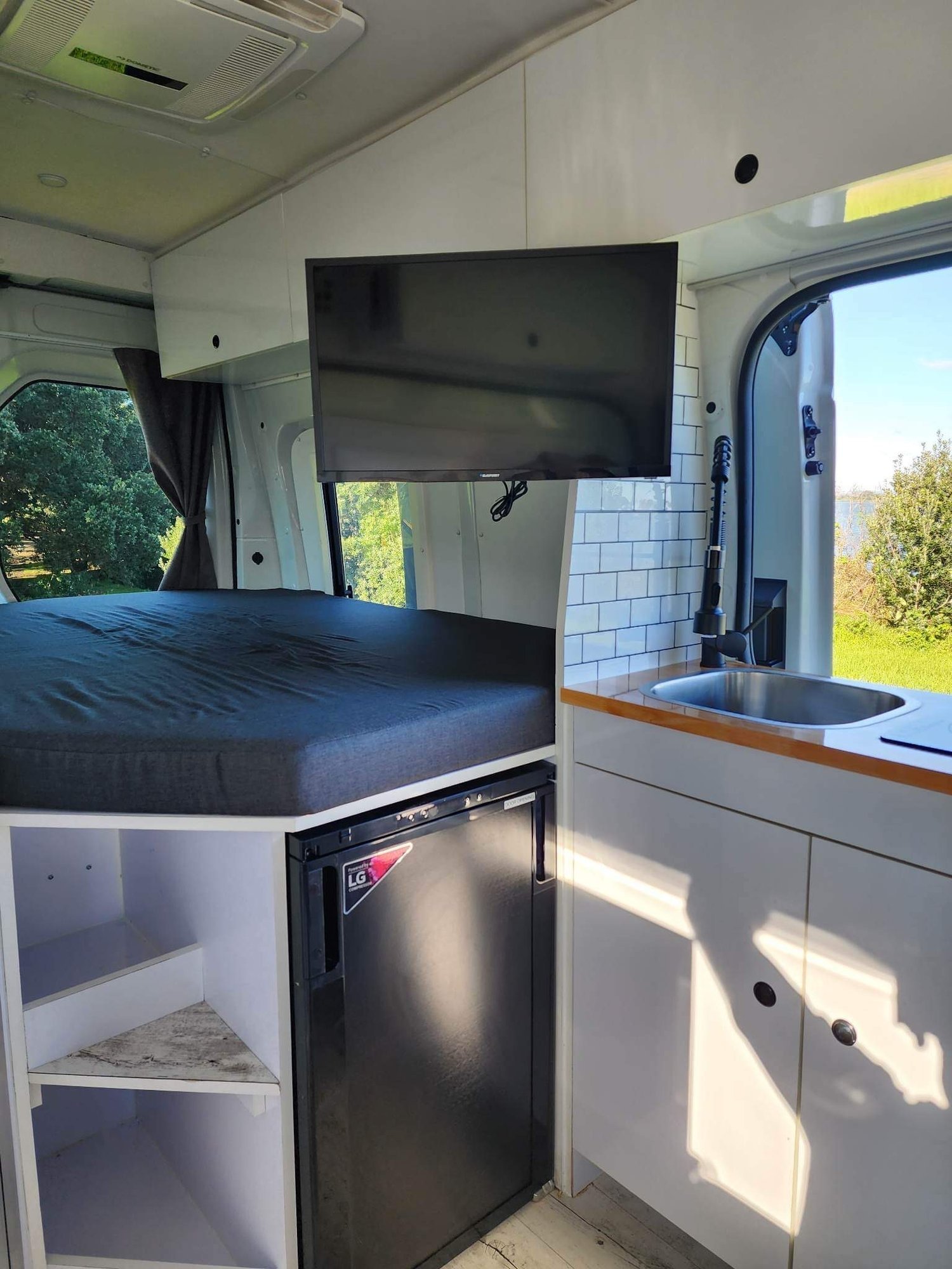Interior of a camper van with a bed, TV, refrigerator, sink, and cabinets.