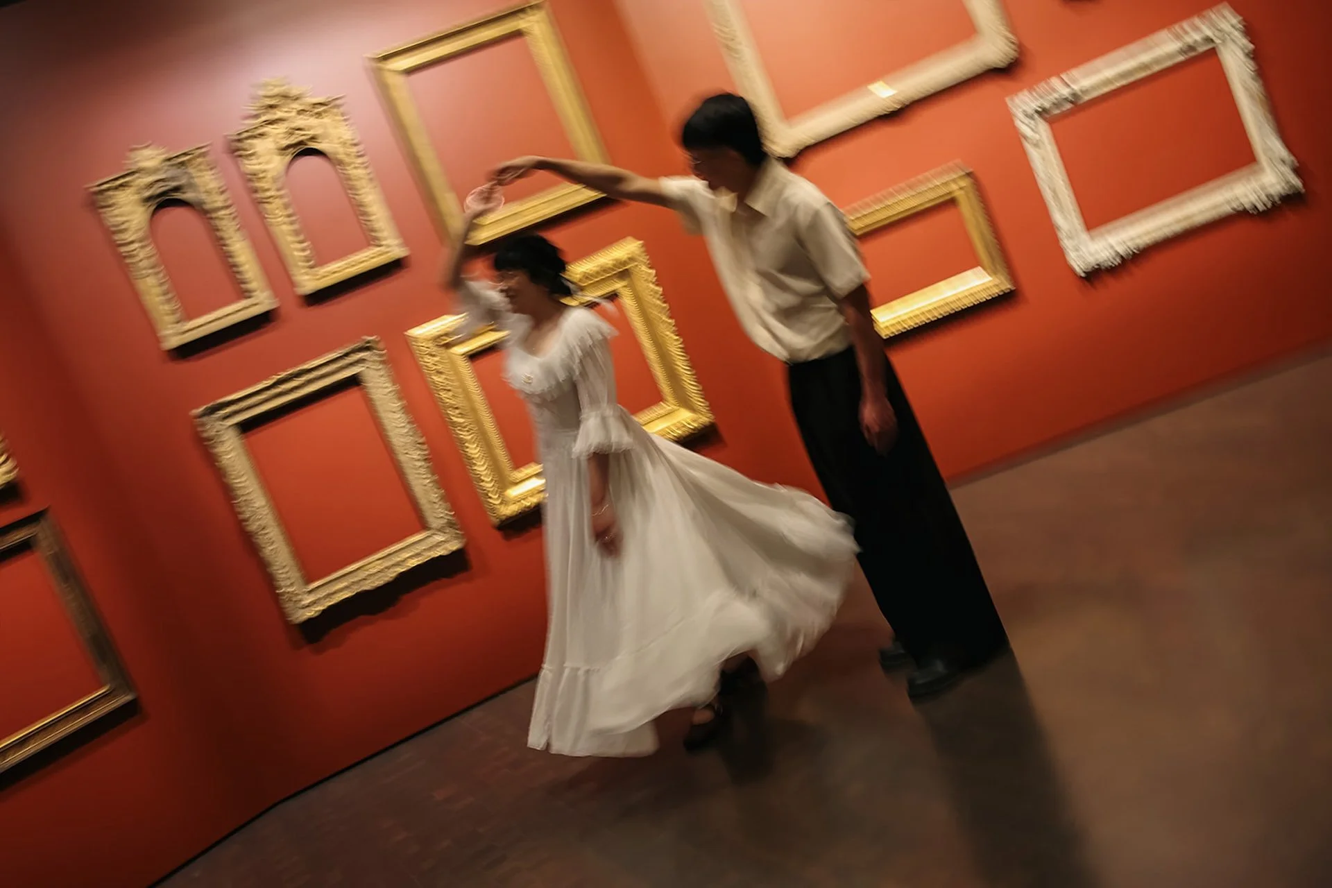 A romantic candid of a couple dancing and twirling together in a gallery with deep red walls, sharing laughter and a quiet moment.