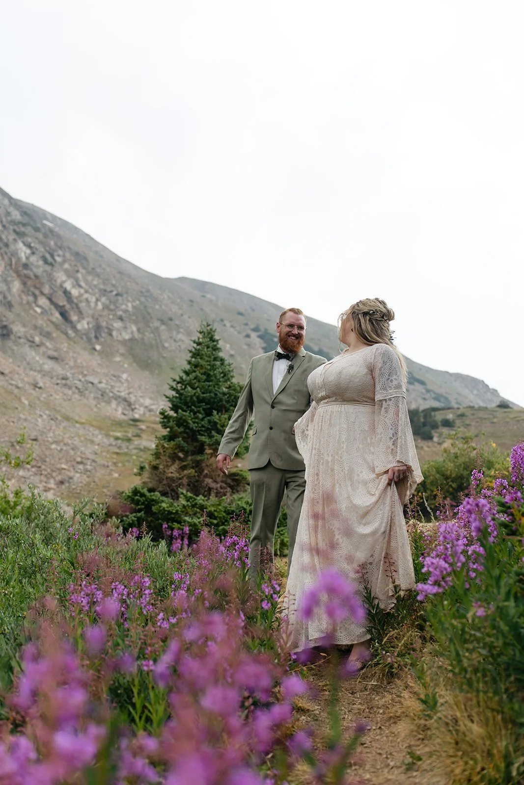 a couple walking through the mountains with wildflowers all around them 