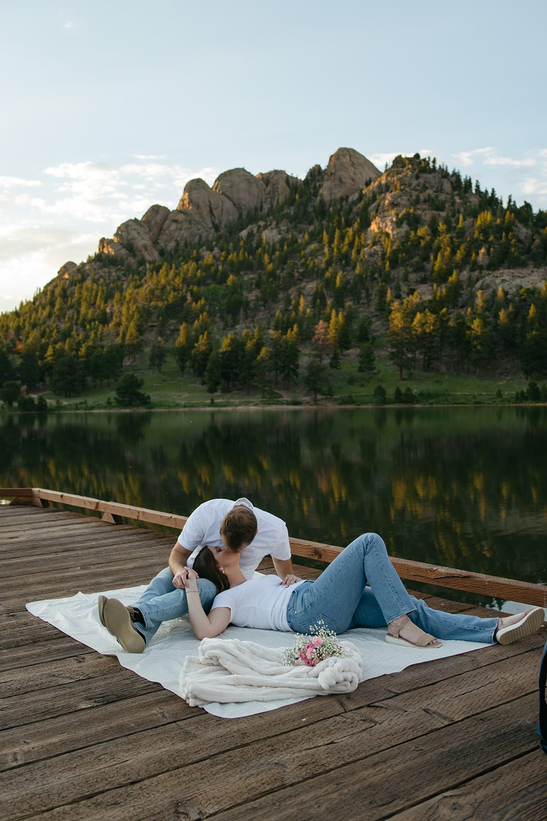 a woman laying in a man's lap as he kisses her with a lake and mountains behind them 