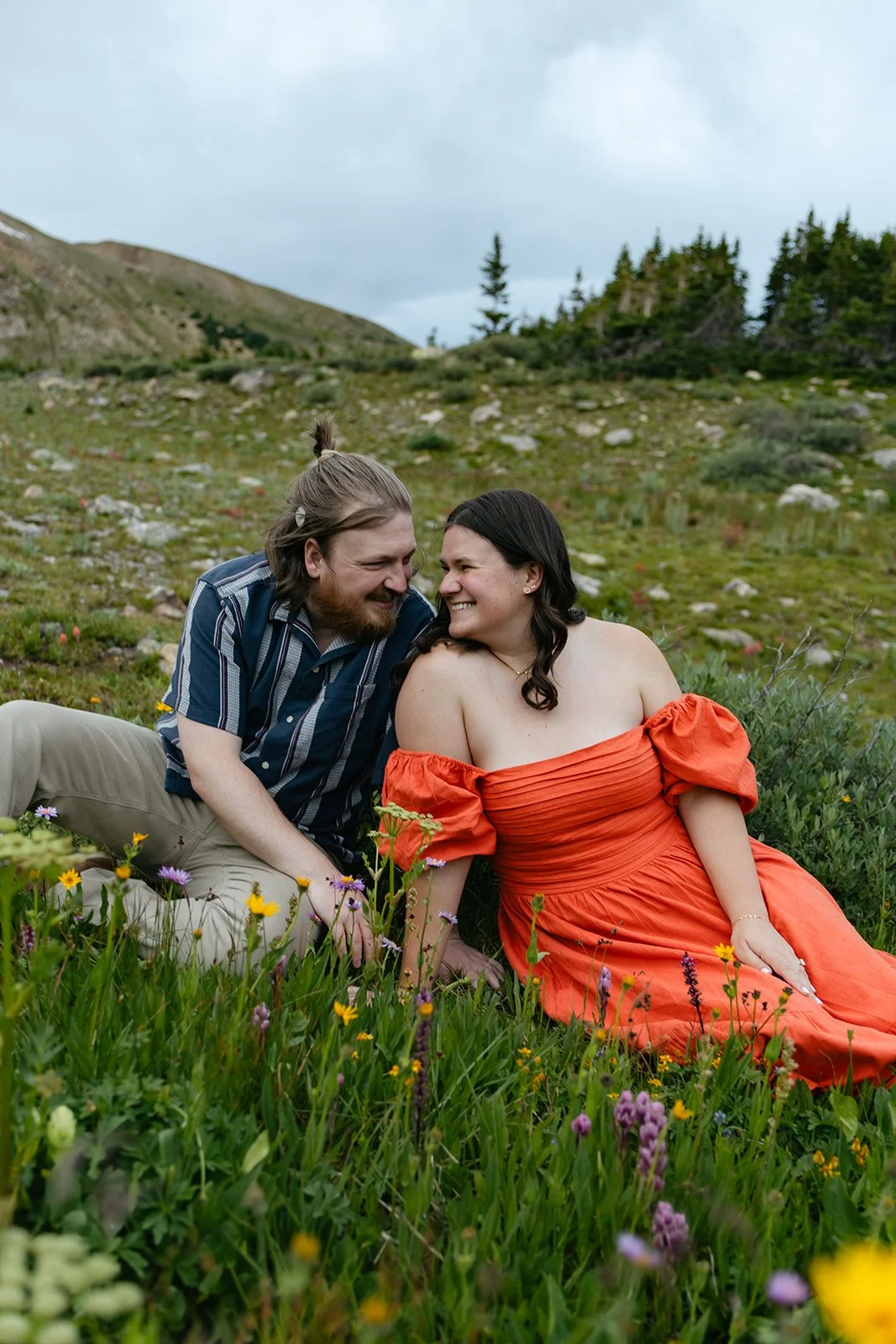 a couple sitting in the mountain wildflowers with pops of color for their Engagement Photo Outfits