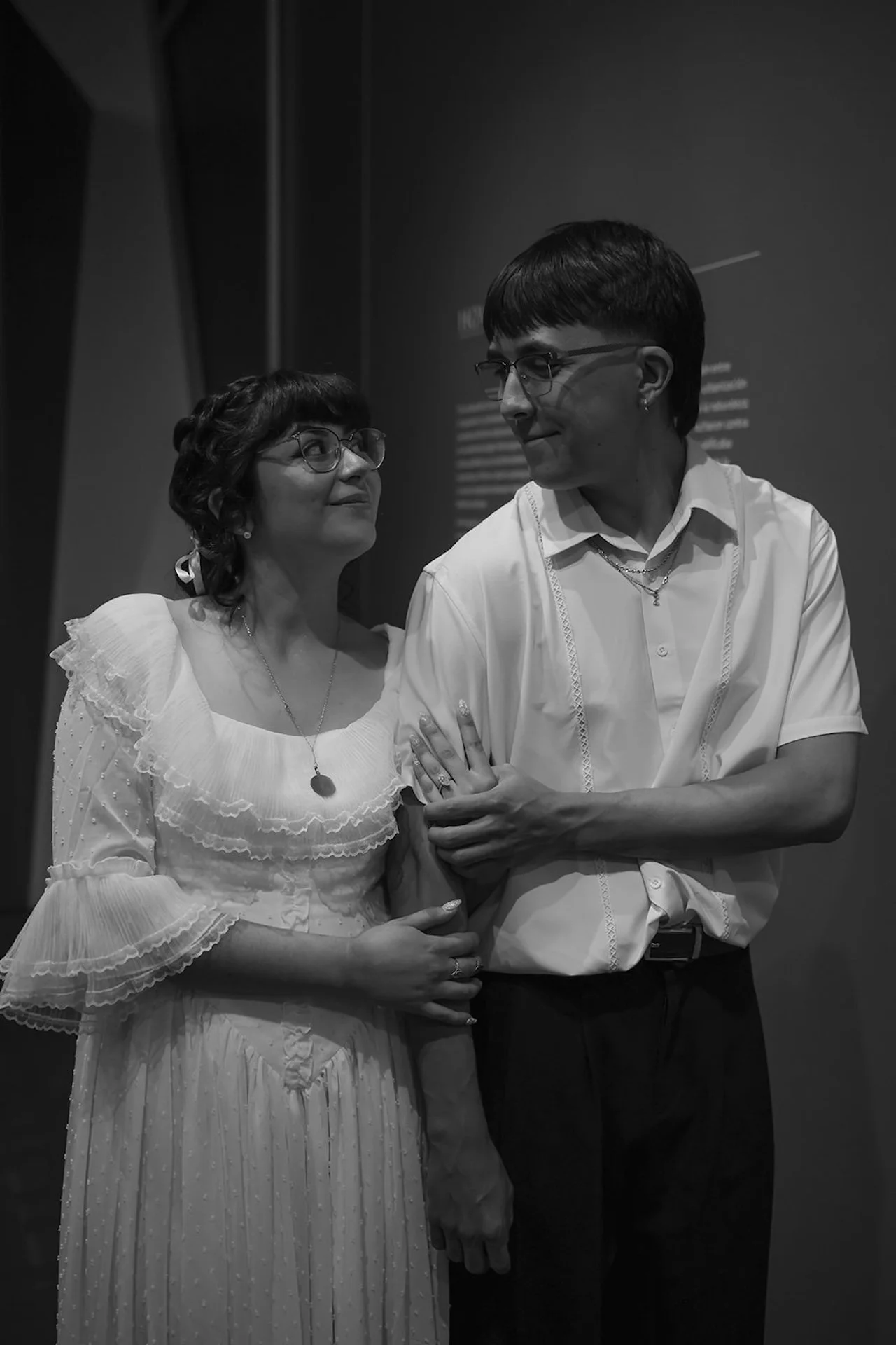 A black and white portrait of a couple walking arm in arm through a museum gallery, sharing a quiet smile as they look at each other.