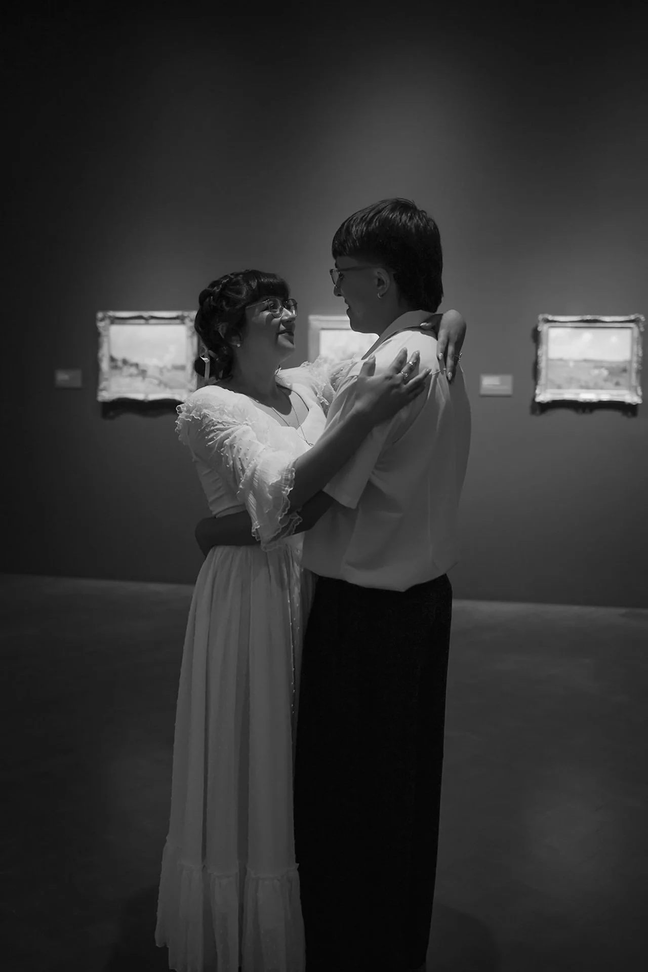 A black and white image of a couple slow dancing in a museum space, framed by artwork in the background for classic, emotional city engagement photos.