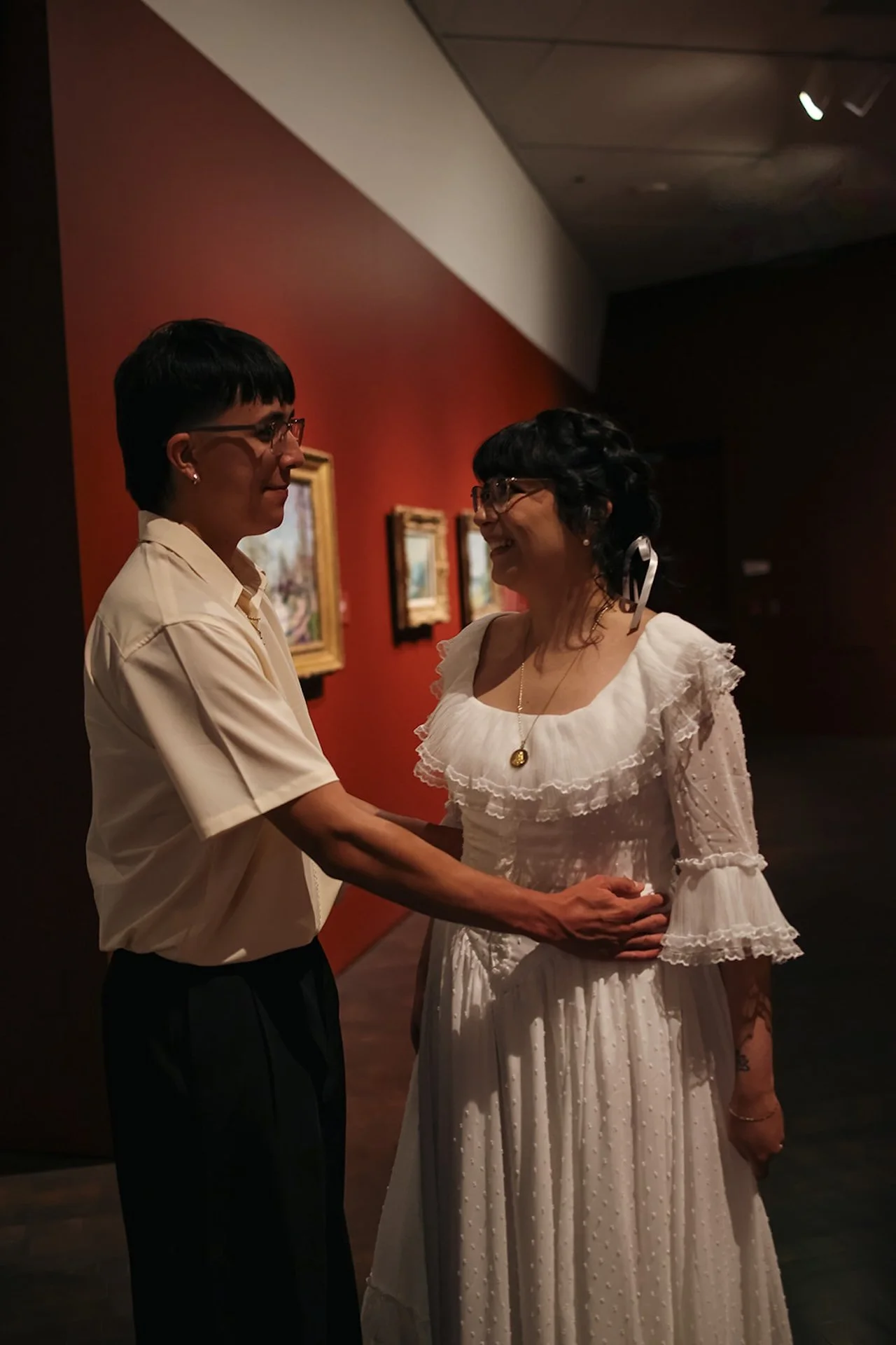 A softly lit portrait of a couple embracing inside a gallery, their faces close as they share an intimate moment surrounded by framed art.