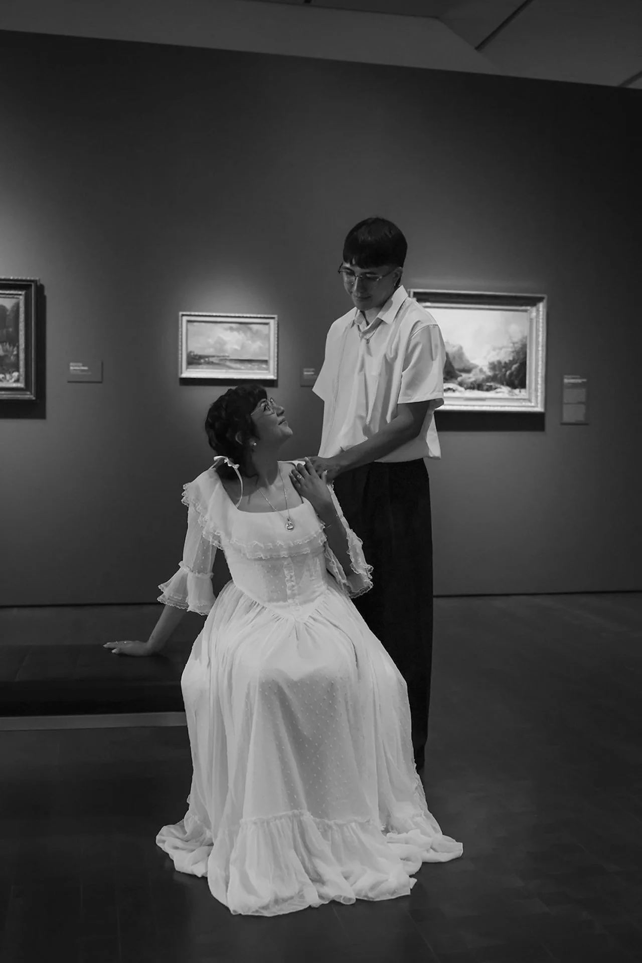 A black and white portrait of a couple seated on a museum bench, holding hands and smiling softly at each other, showcasing elegant city engagement photos indoors.