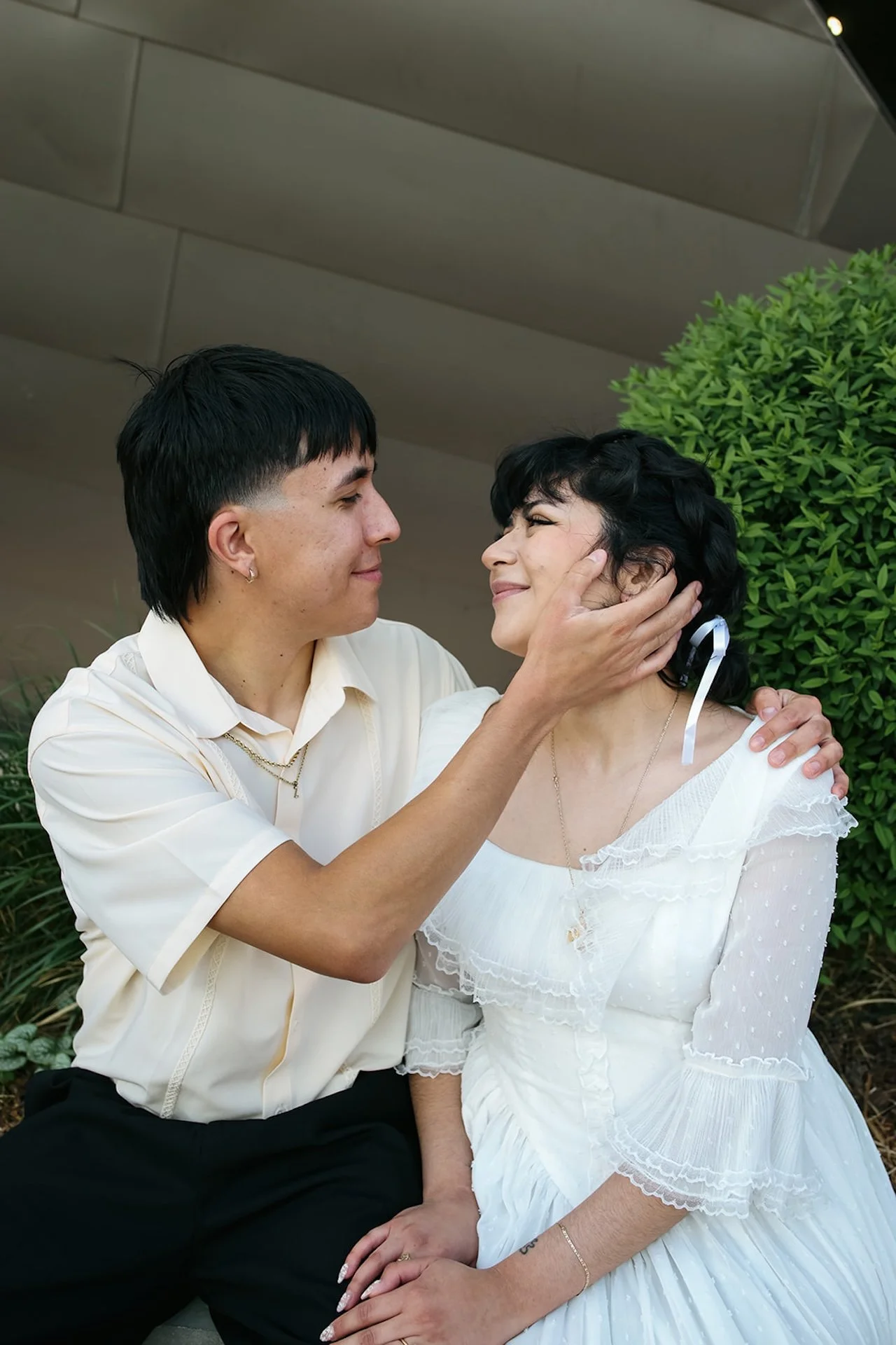A close-up portrait of a couple gazing at each other lovingly, with one partner gently holding the other’s face while seated outdoors near a museum wall.