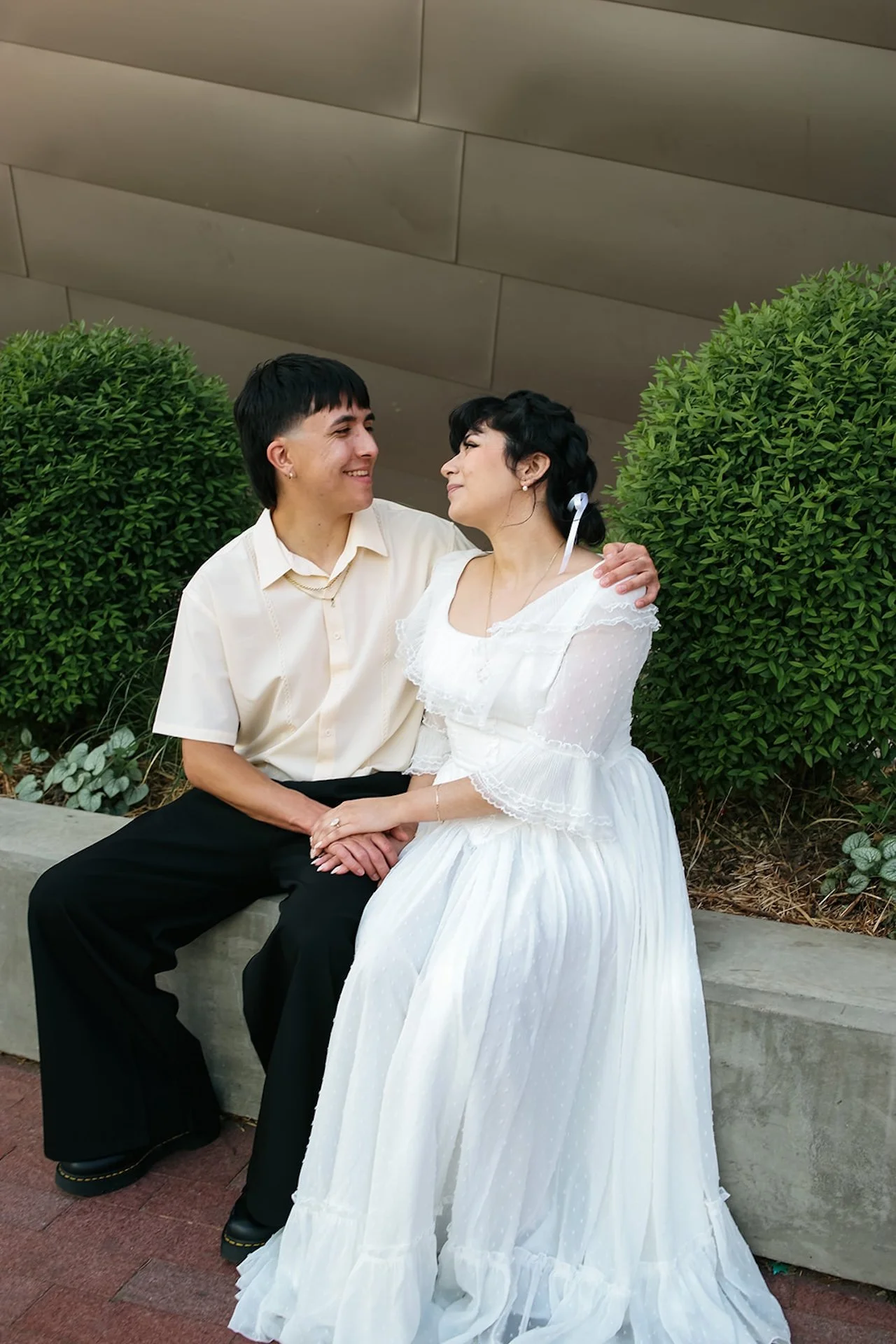 A candid moment of a couple sitting close together on a low concrete ledge, smiling at each other with manicured greenery and modern architecture behind them, perfect for city engagement photos.