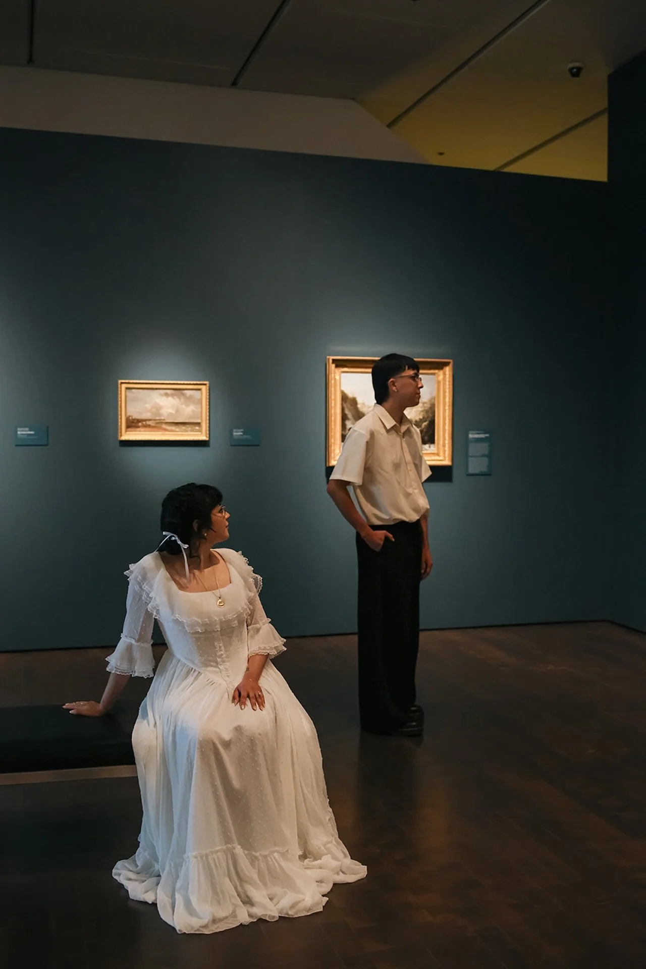 A wide shot of a woman seated on a museum bench while her partner stands nearby, both surrounded by classic artwork in a calm gallery space.