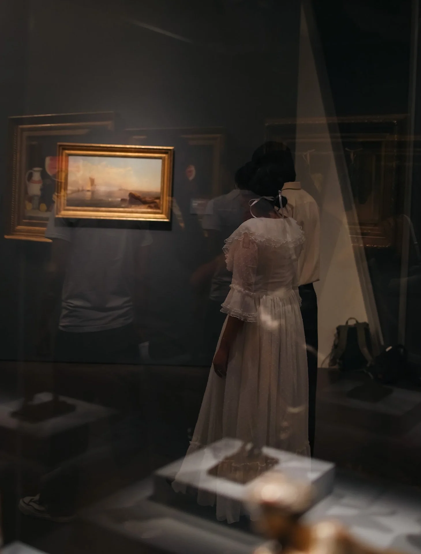 A layered reflection shot of a couple viewed through glass cases, with artwork and visitors softly blending into the scene.